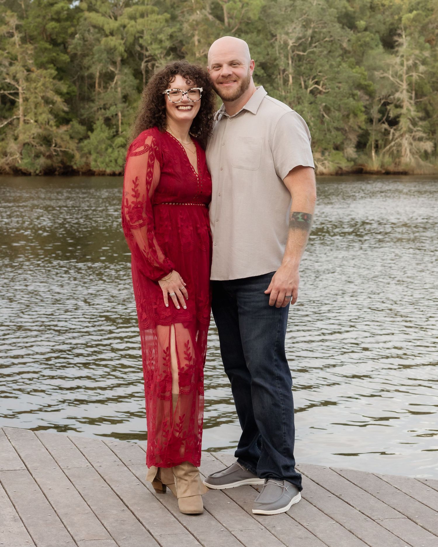 Smiling couple posing on a dock by a lake, woman in red lace dress and man in beige shirt.