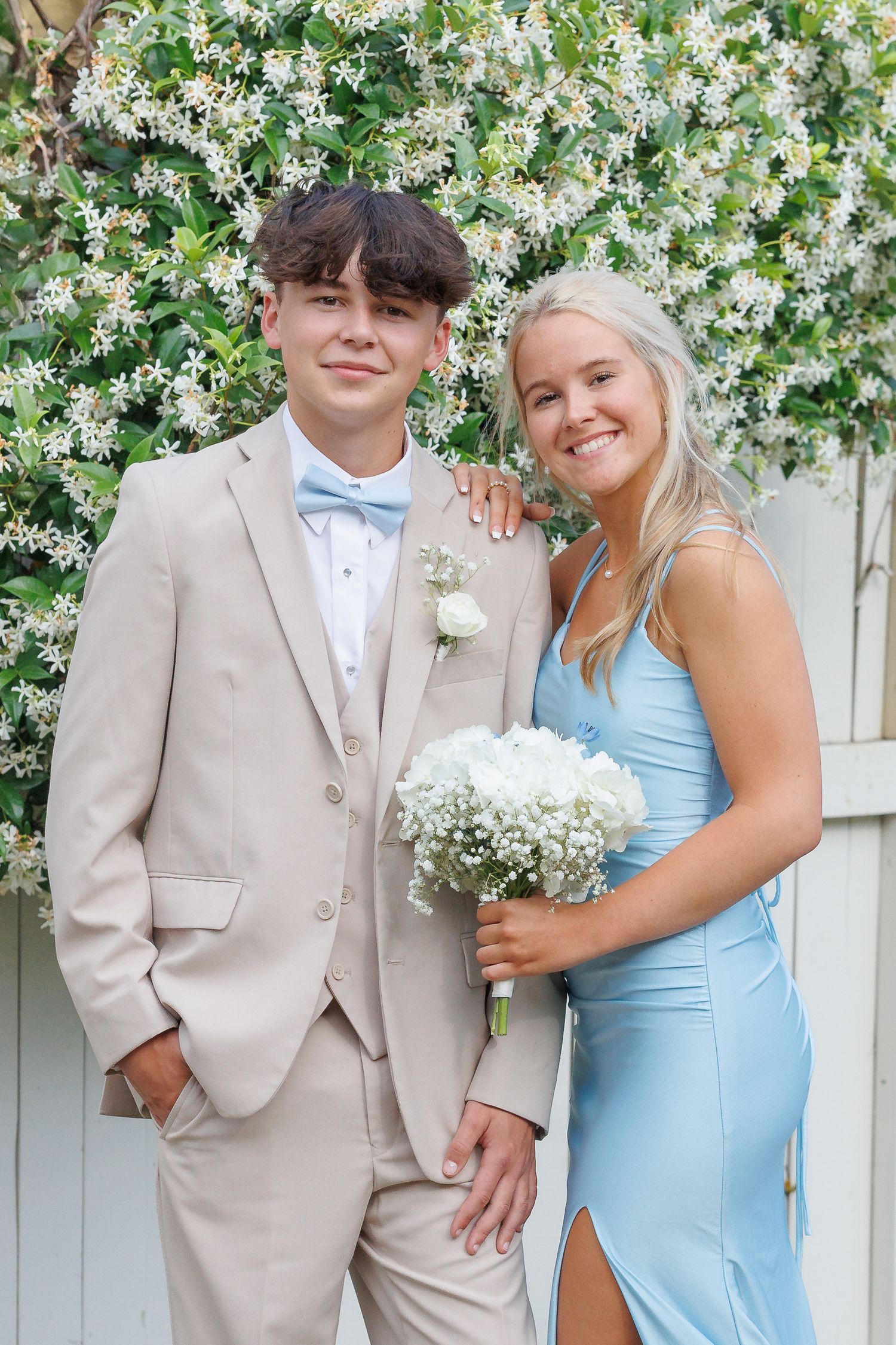 A nicely dressed couple poses against a white jasmine flower wall for a formal event photo.
