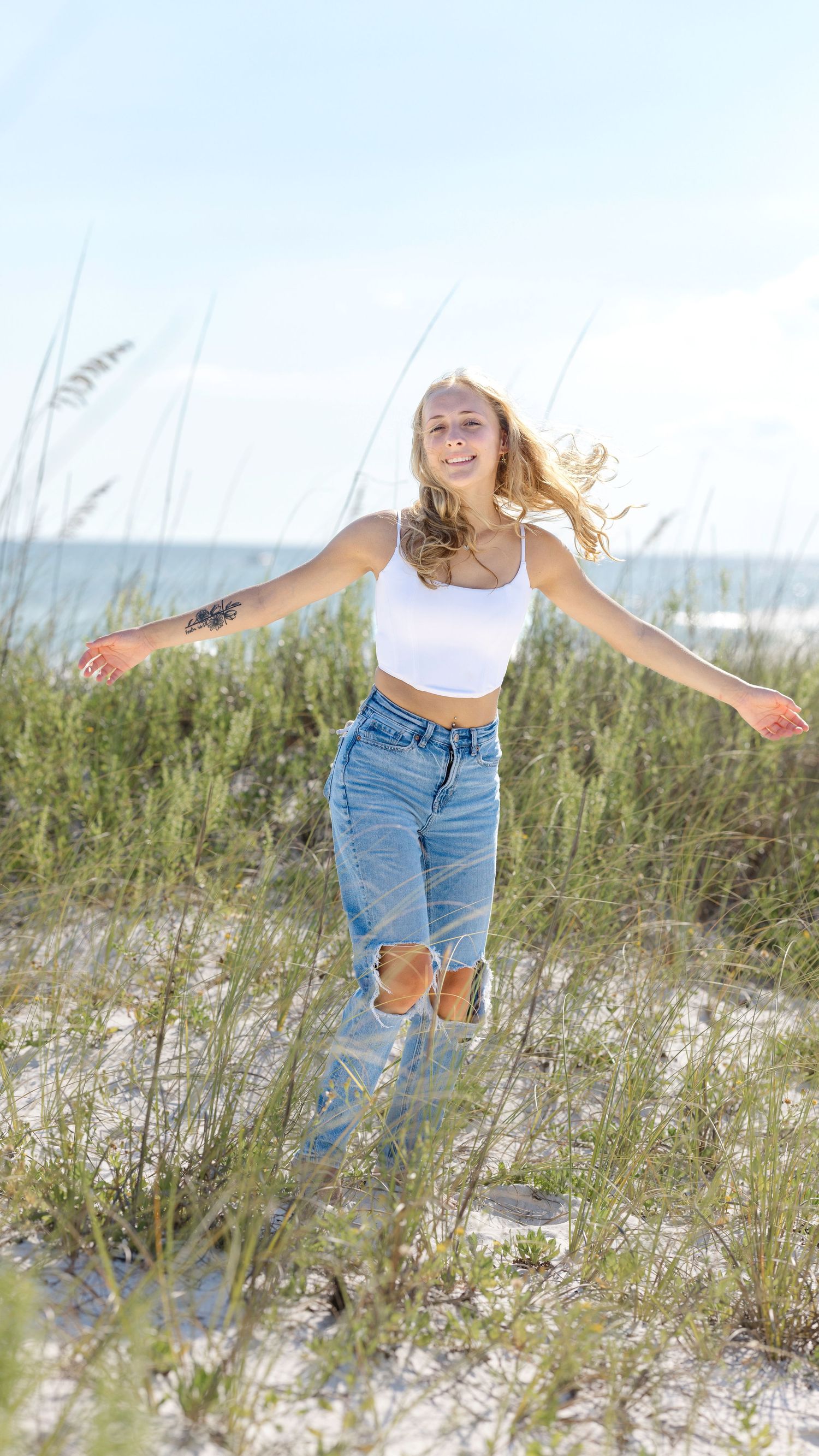 Person in casual summer outfit jumping joyfully on a sandy beach with beach grass and ocean in background.