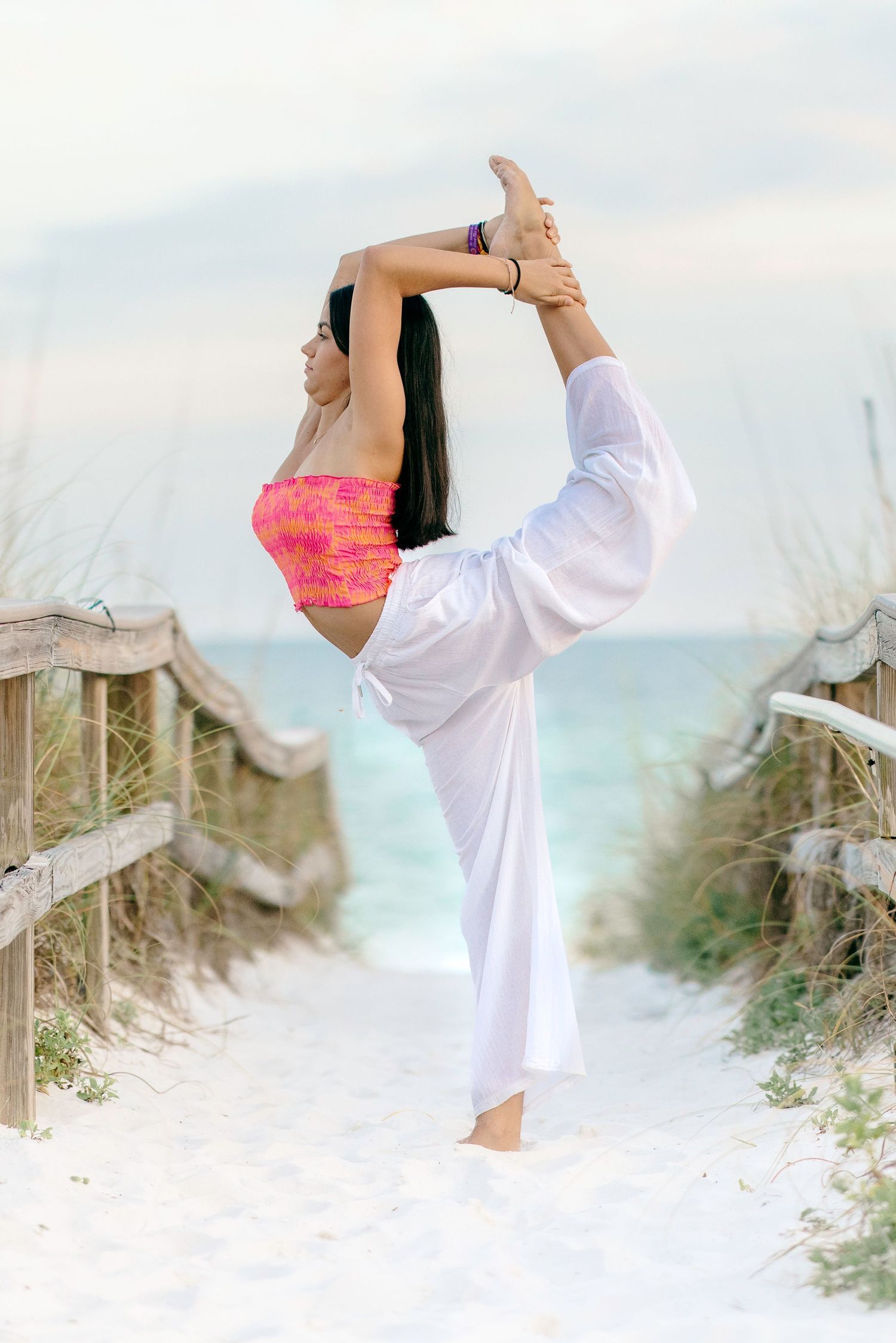 Woman in white kimono performing standing leg stretches on sandy dunes against scenic beach backdrop.
