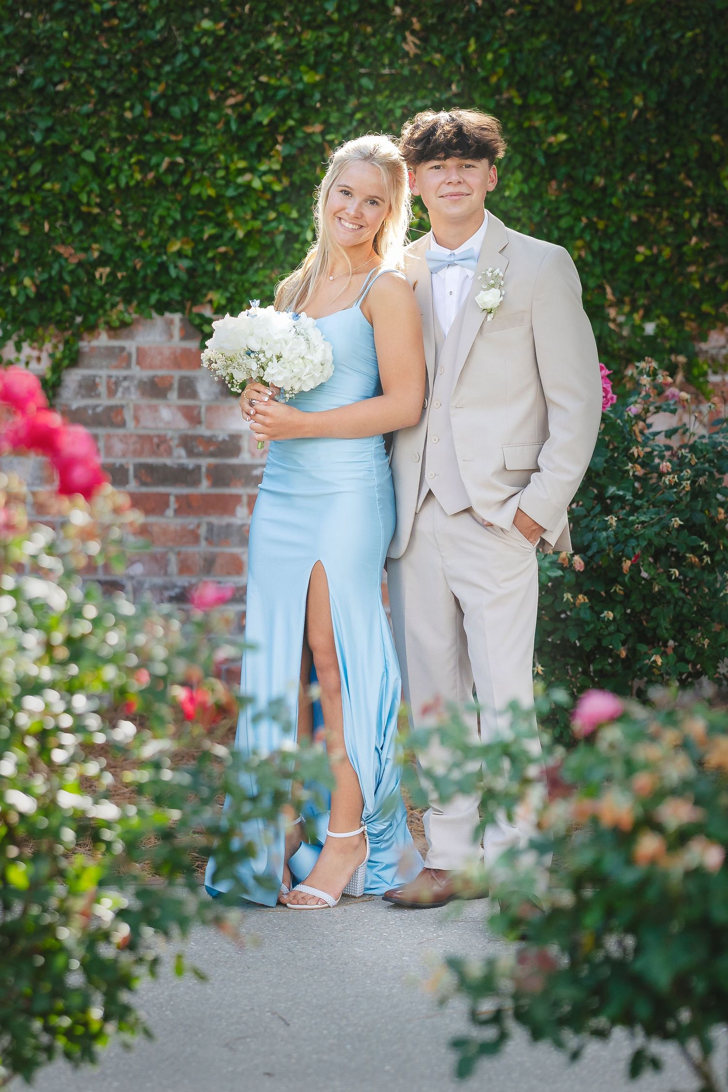 A couple in light blue dress and beige suit poses in front of a garden wall with blooming roses and greenery.