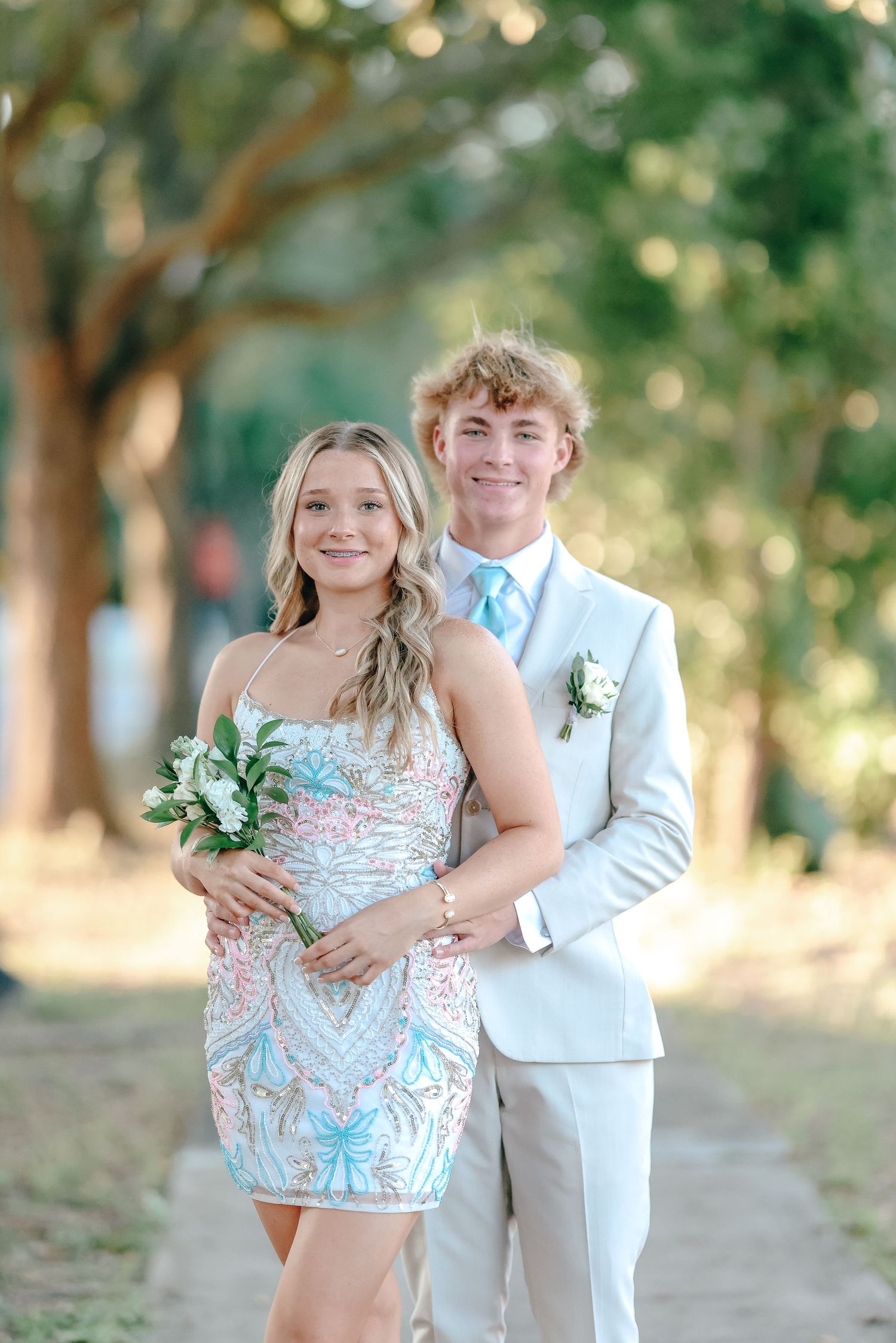 A couple poses outdoors for a formal event photo in light colored attire with a bouquet against a natural backdrop.