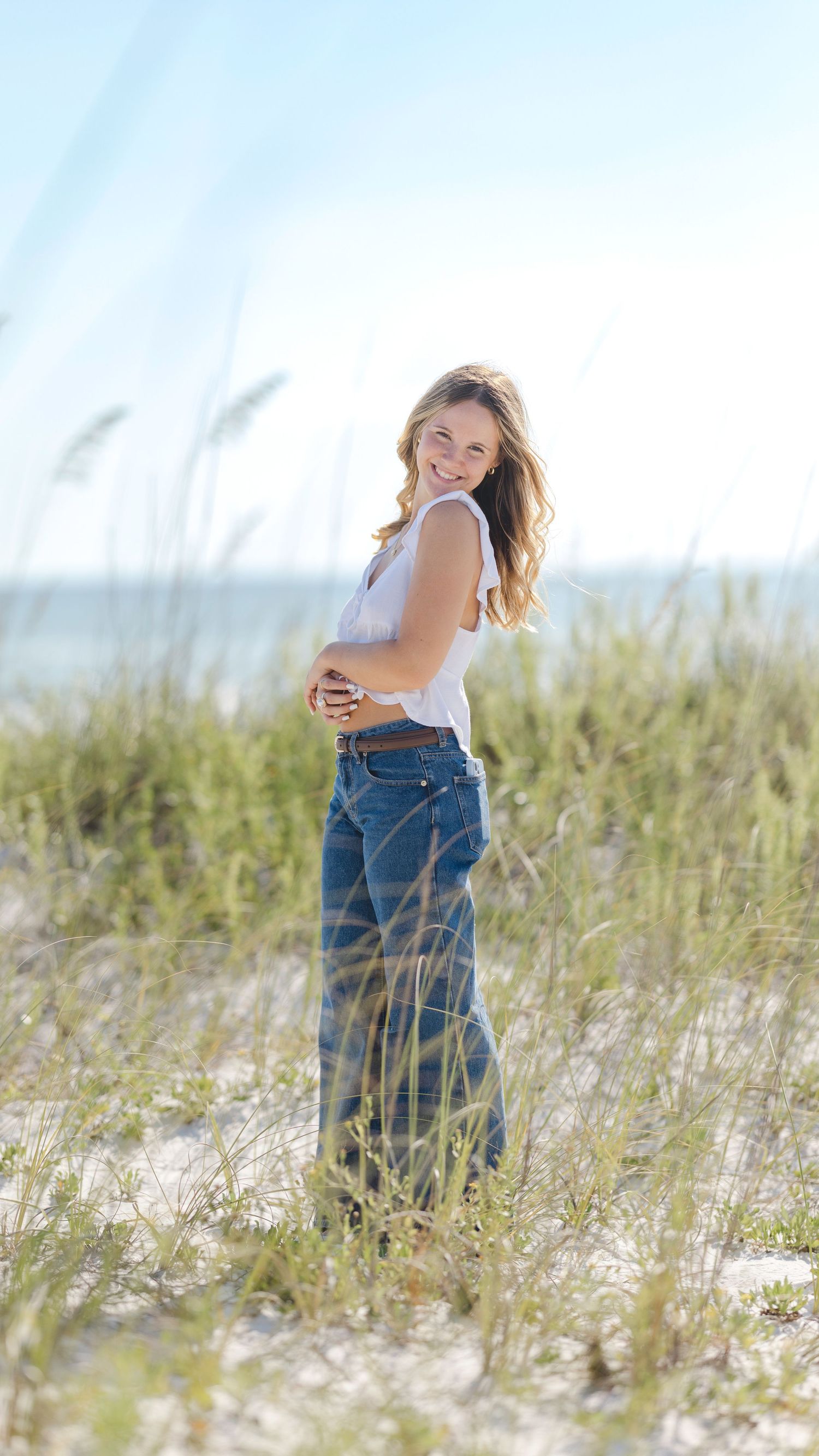 A person in a white tank top and jeans stands smiling in beach grass on a sunny day by the ocean.