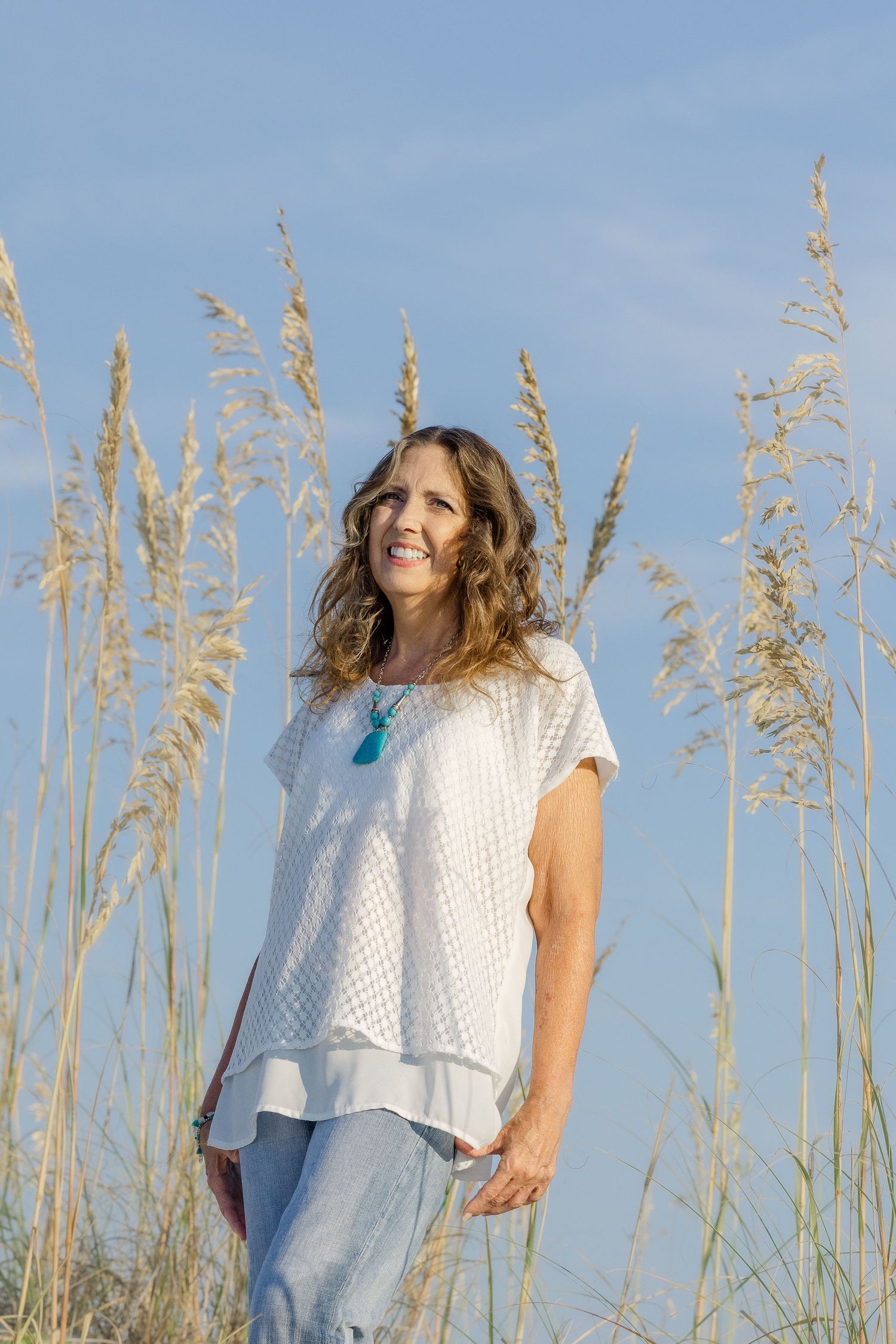 A person in a white eyelet top poses joyfully among tall sea oats against a bright blue sky.