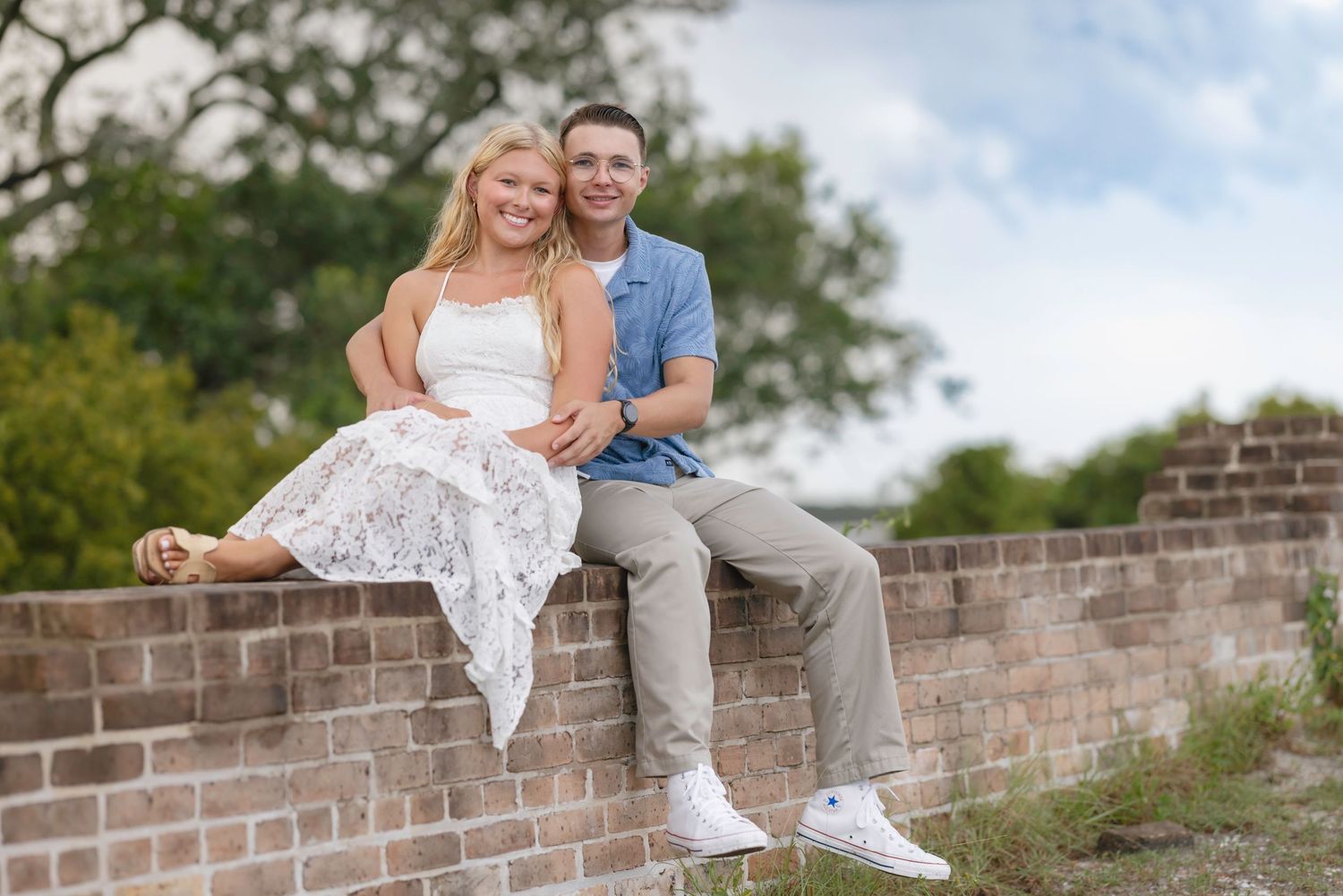 Happy young couple sitting together on a rustic brick wall outdoors, smiling with trees and sky in background.