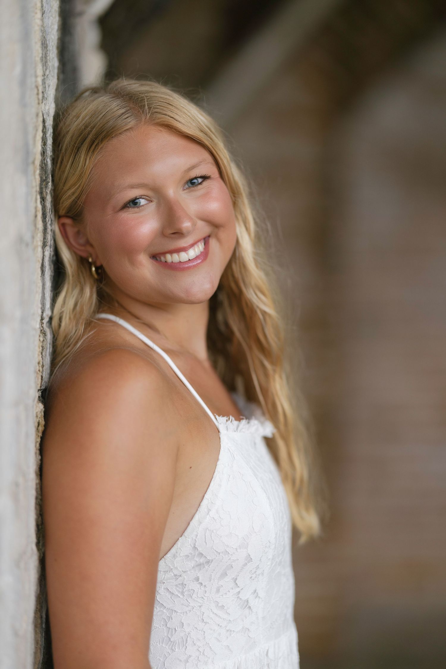 Smiling blonde teenage girl in white lace dress leaning against a stone wall outdoors.