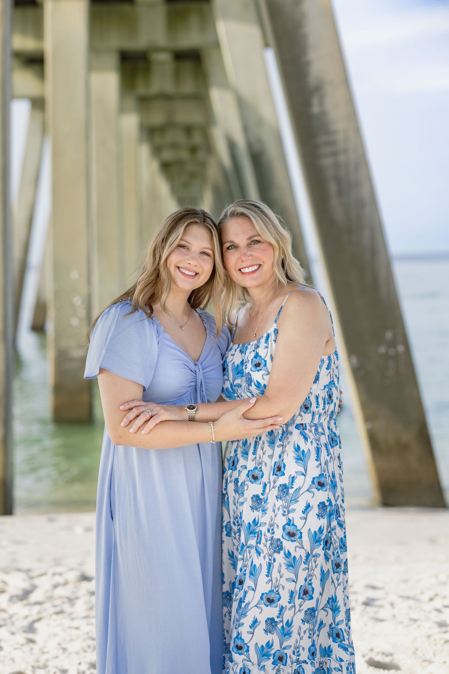 Two smiling women in blue dresses pose together on a sandy beach beneath a wooden pier.