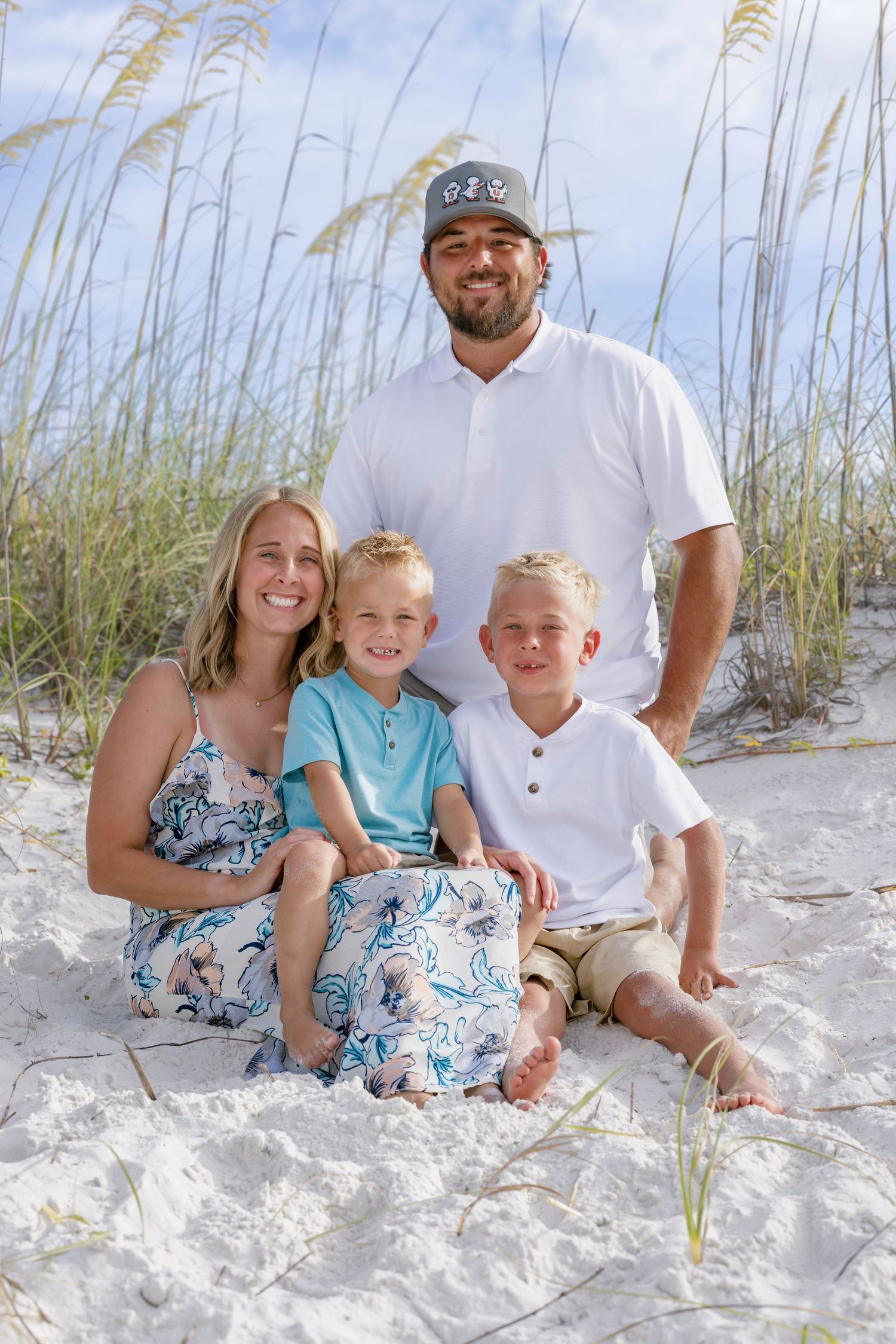 Family of four poses on white sand beach dunes with sea oats, mom in floral dress, dad in white polo, two blonde boys.