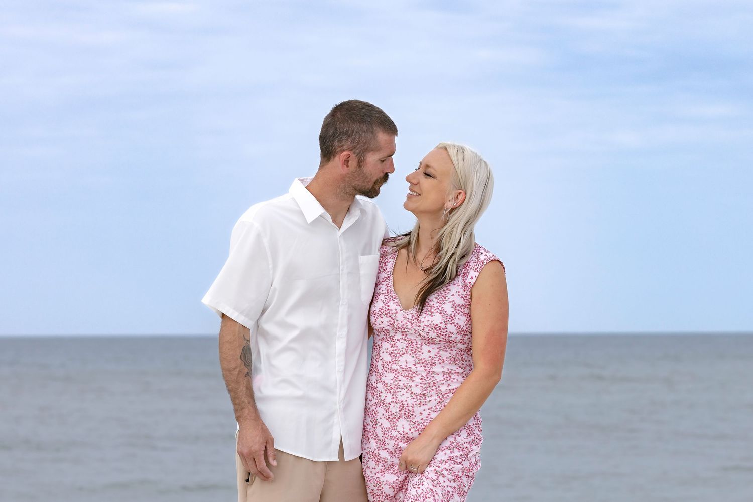 Couple smiling at each other on a beach, man in white shirt and woman in pink floral dress with ocean backdrop.
