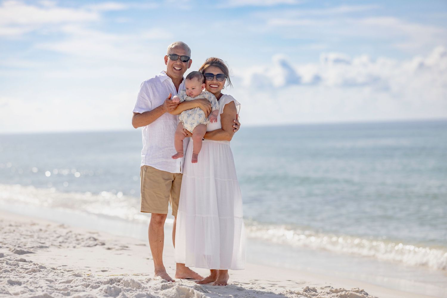 A happy family of three poses on a white sandy beach, with parents holding their baby, wearing white outfits on a sunny day.