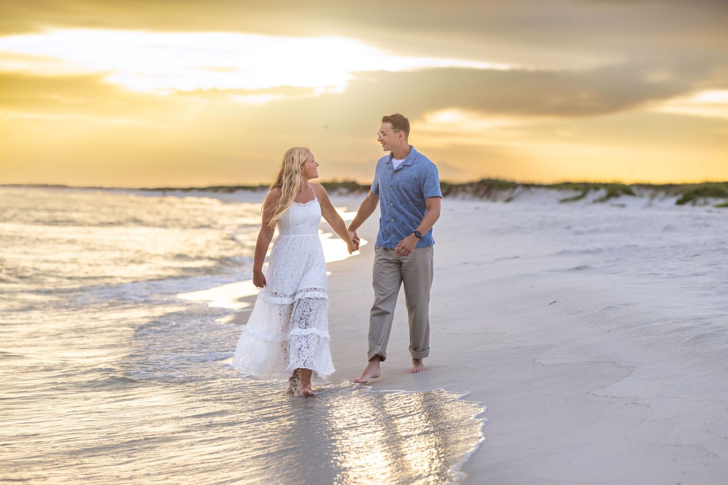 Couple holding hands walking on a sandy beach at golden sunset, woman in white dress, man in casual gray pants.