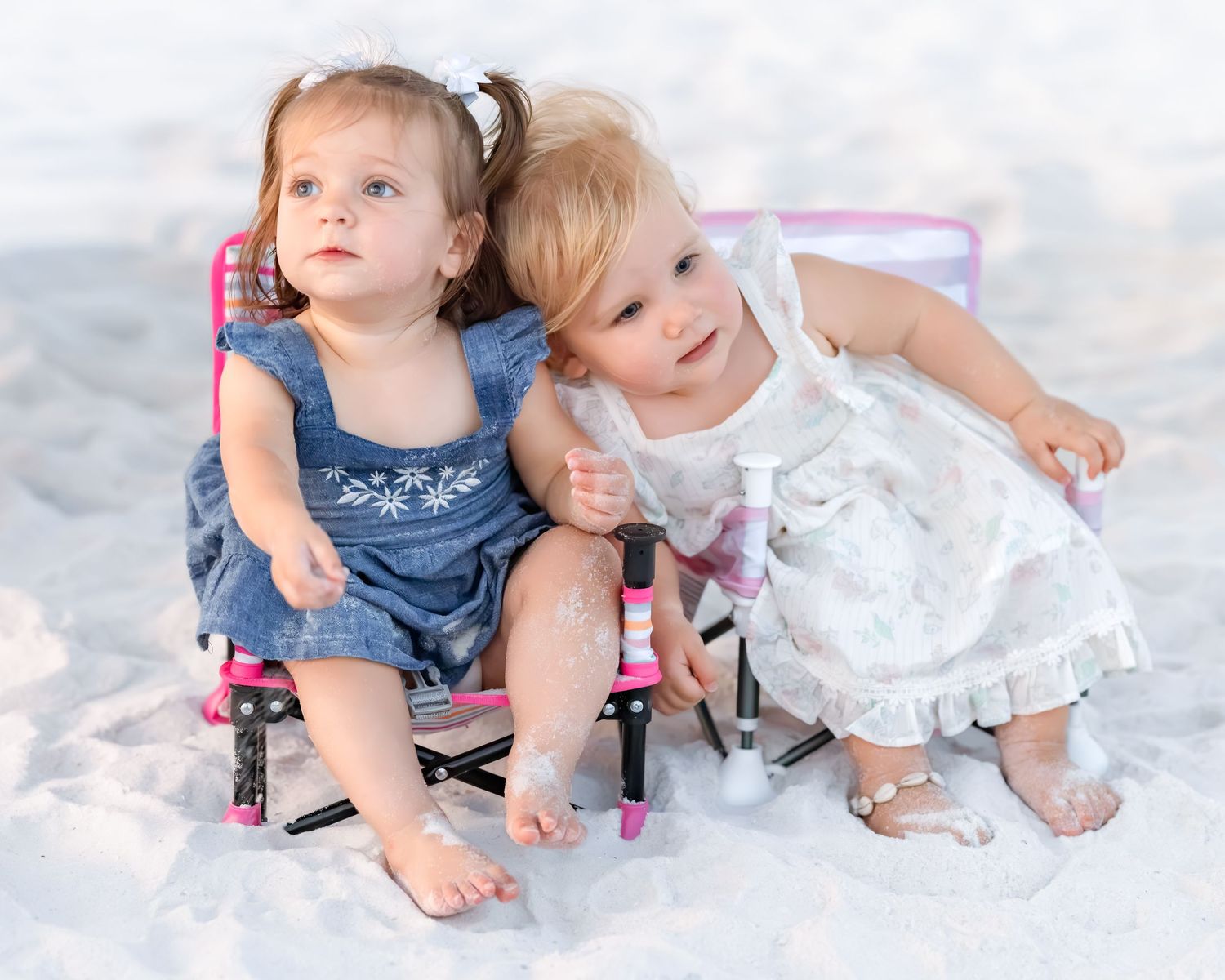 Two adorable toddler girls sit together on white sand beach, one in denim dress and one in white ruffled dress.