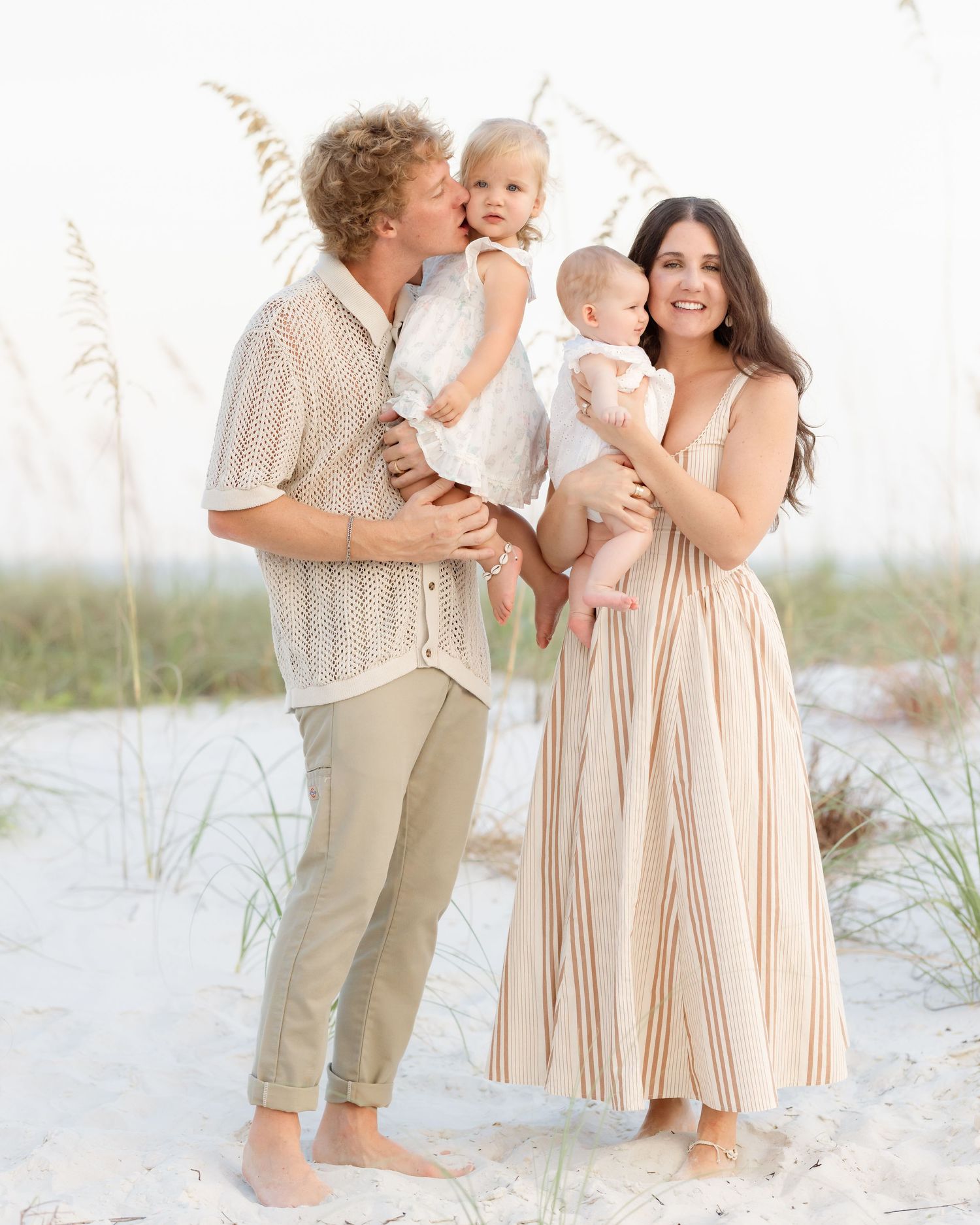 Young family of four at beach, curly-haired dad kissing toddler while mom holds infant in striped maxi dress.