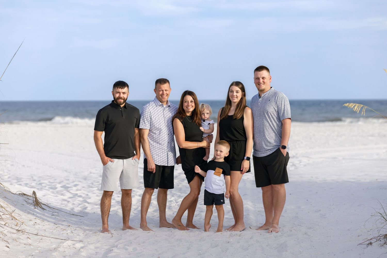 Family of six posing together on a white sand beach with ocean waves in the background on a sunny day.