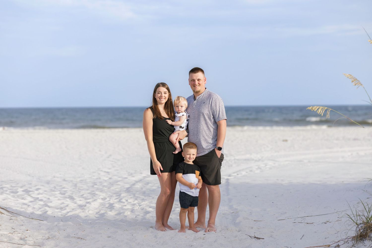 Family of four posing on a white sand beach, parents with two young children smiling with ocean in background.