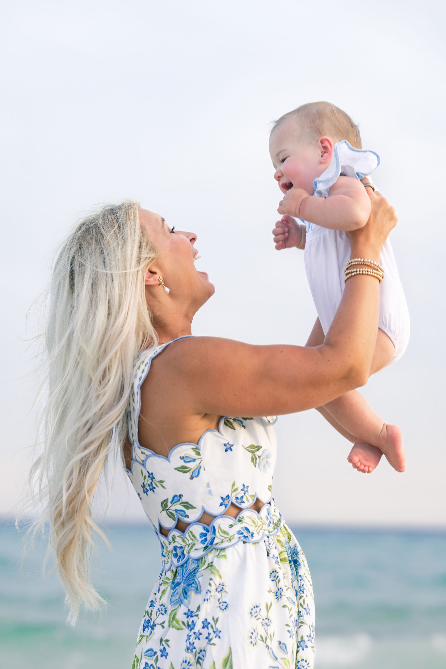 Blonde woman in floral dress lifting happy baby at beach.