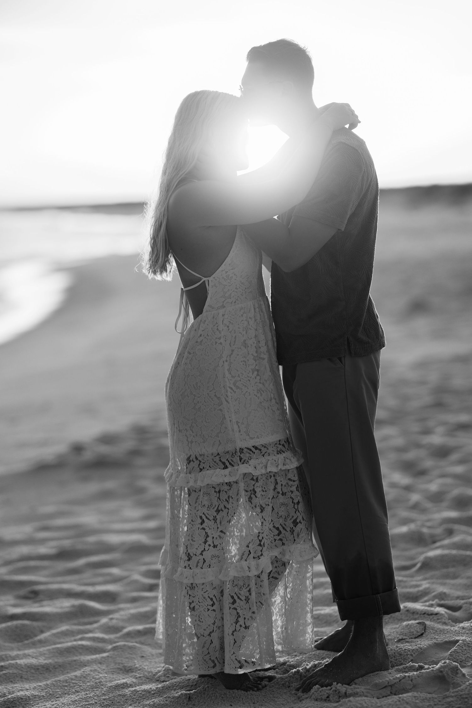 Black and white photo of a couple embracing on a beach at sunset, sun flare glowing between them.