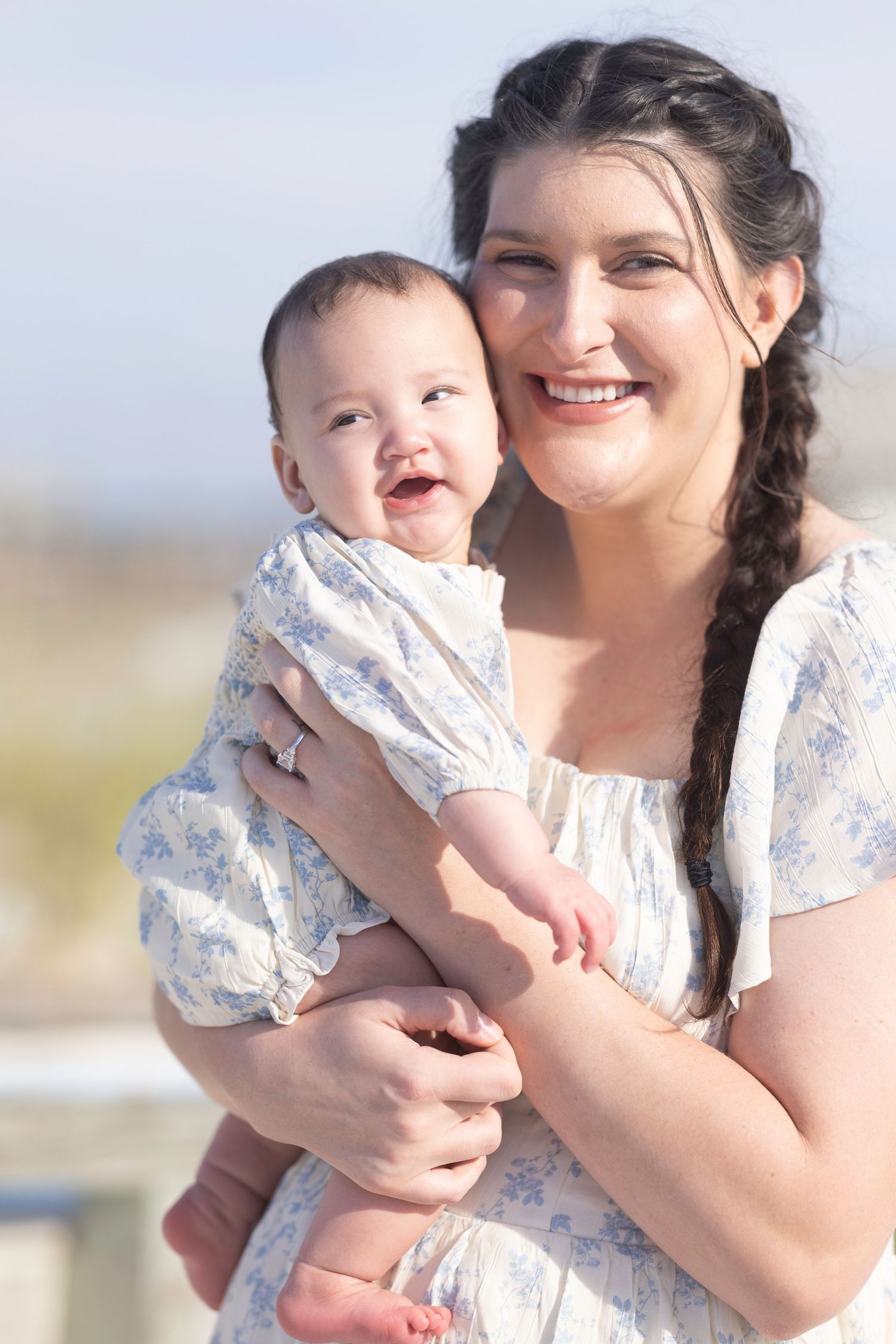 Smiling mother with braided hair holds her happy laughing baby outdoors in matching floral outfits.