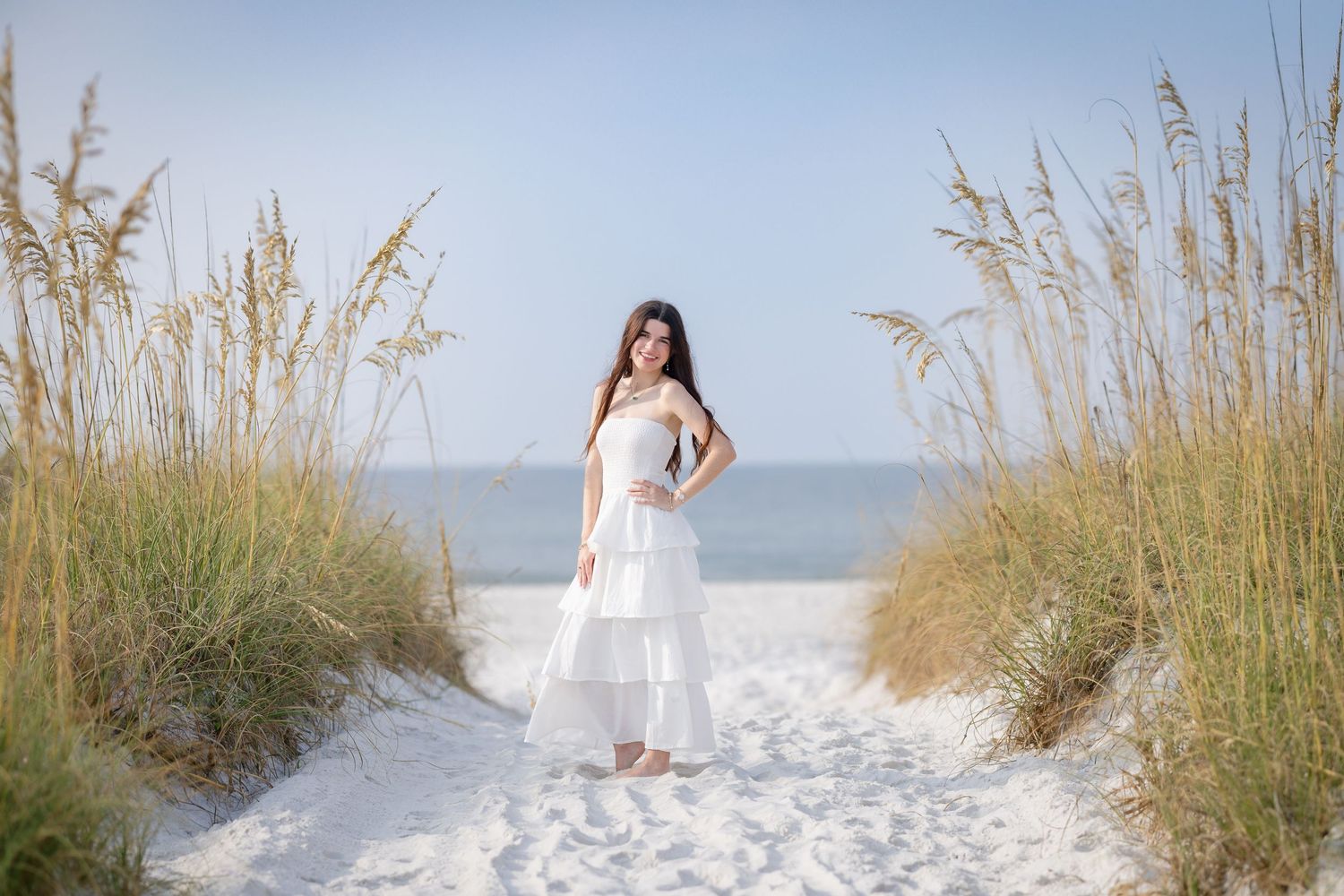 Young woman in white dress standing on sandy beach path flanked by sea oats with ocean horizon in background.