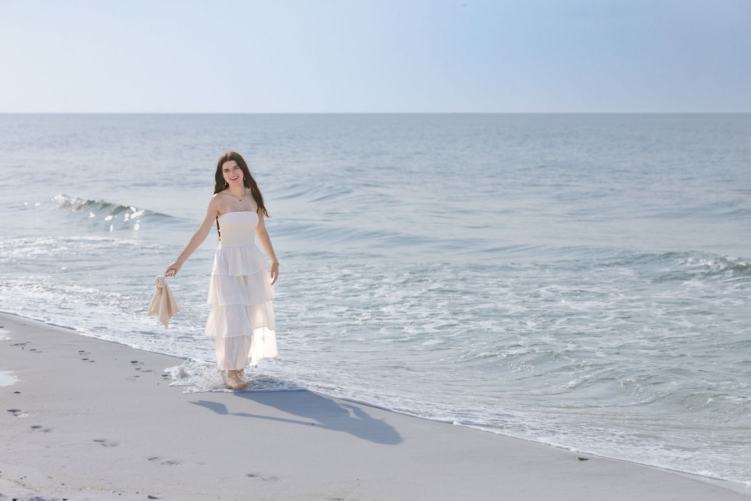 Young woman in white maxi dress walking along sunny ocean shoreline carrying a bag.