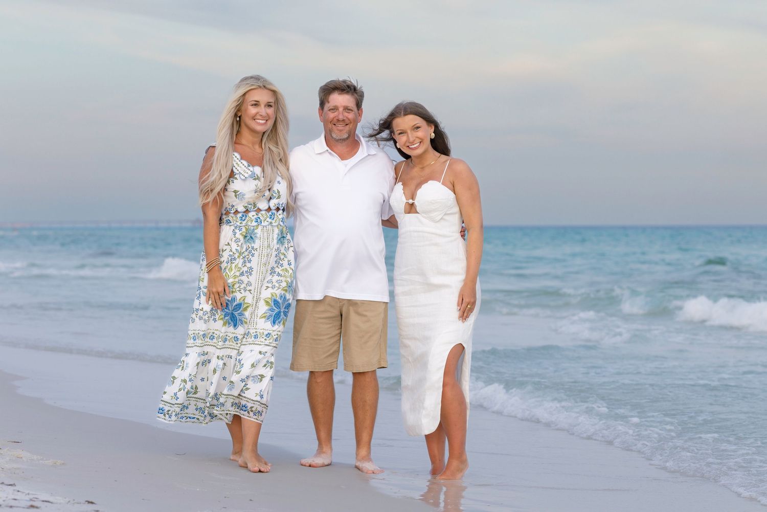 A man and two women smiling together on a sunny beach with ocean waves in the background.