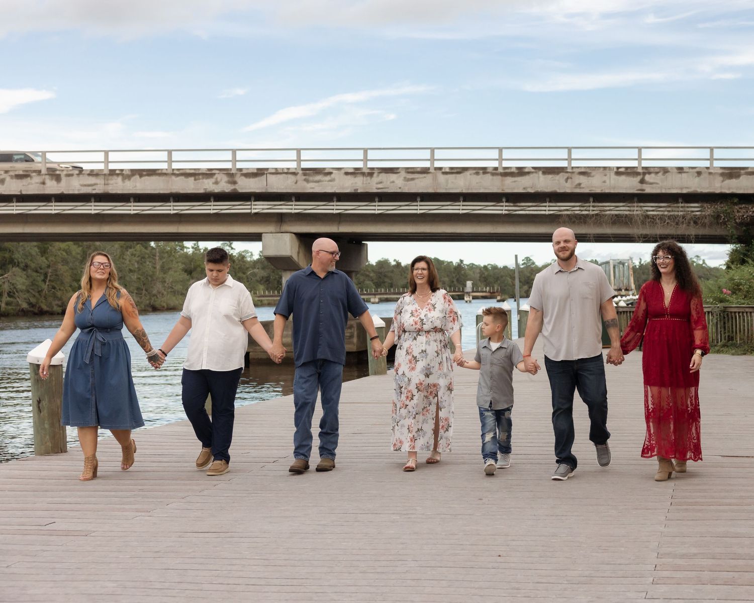Family of seven holding hands walking on a waterfront dock with a bridge in the background on a sunny day.