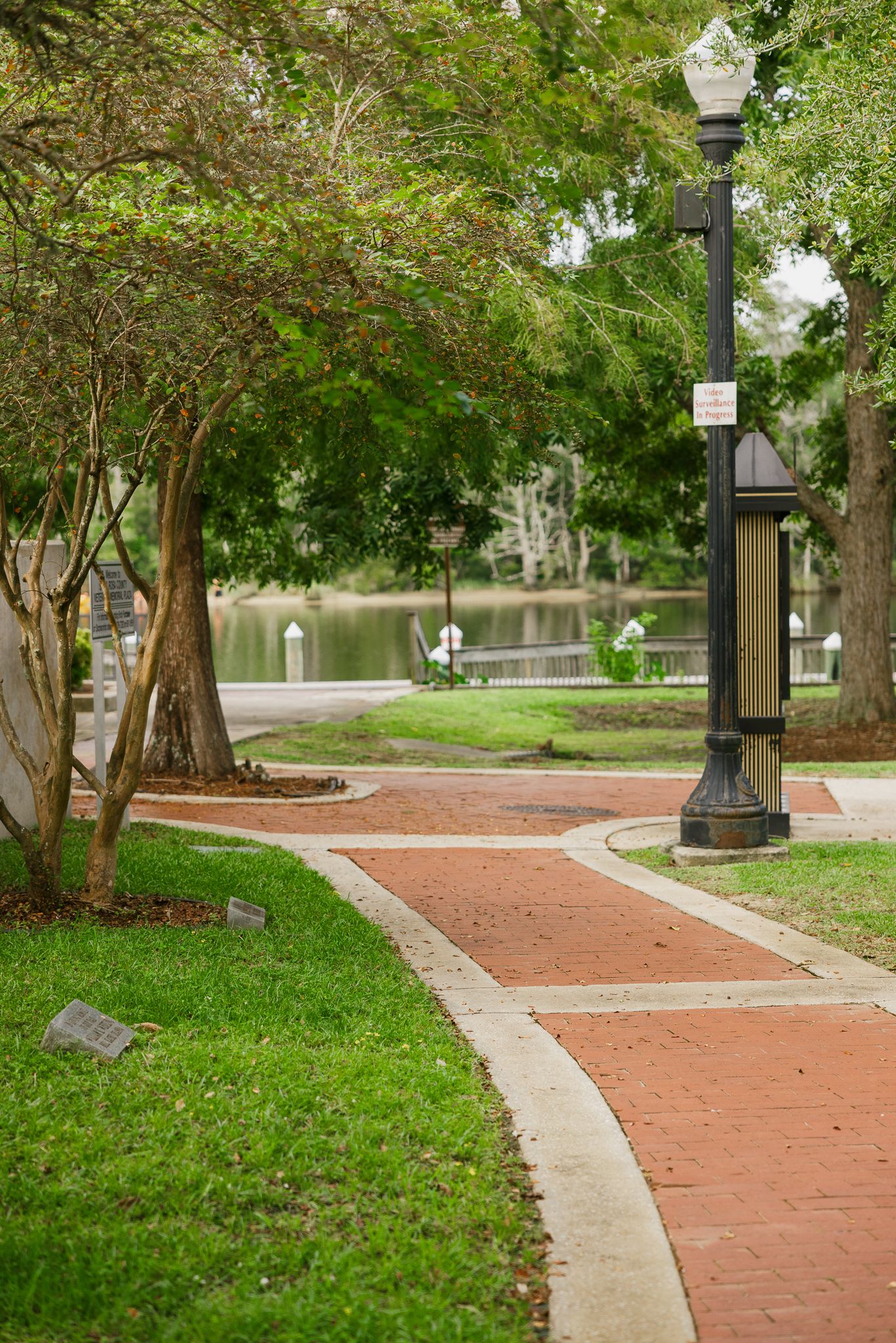 A winding brick pathway through a lush green park with a decorative lamp post and serene pond in the background.