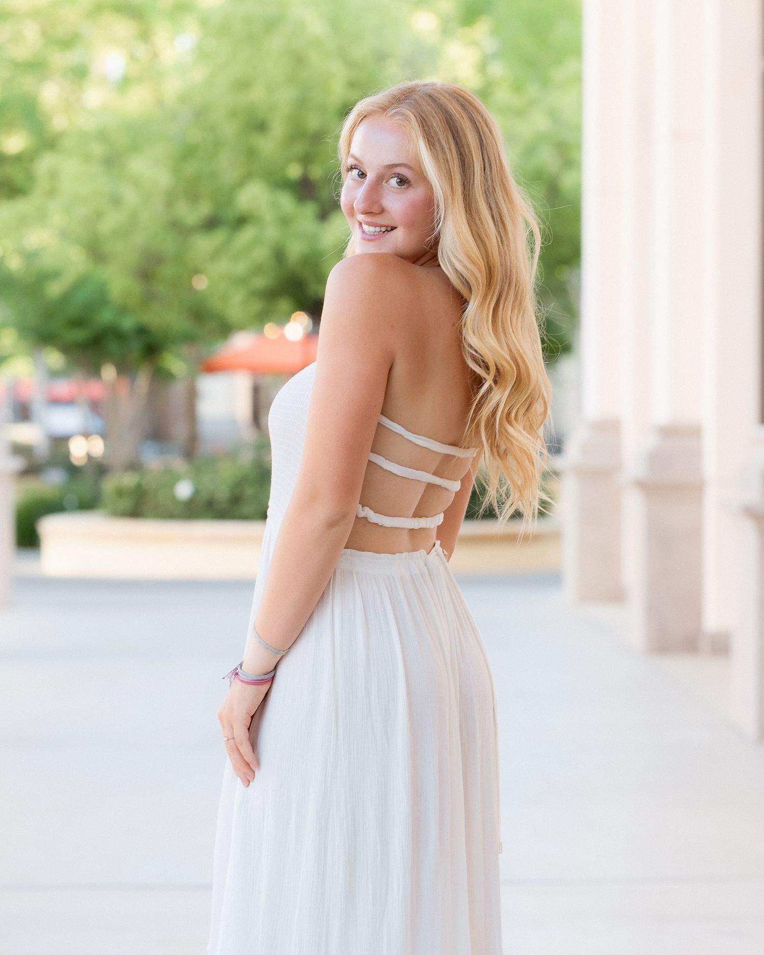 Young woman in white backless gown with strappy detail, long blonde wavy hair, smiling over shoulder outdoors.