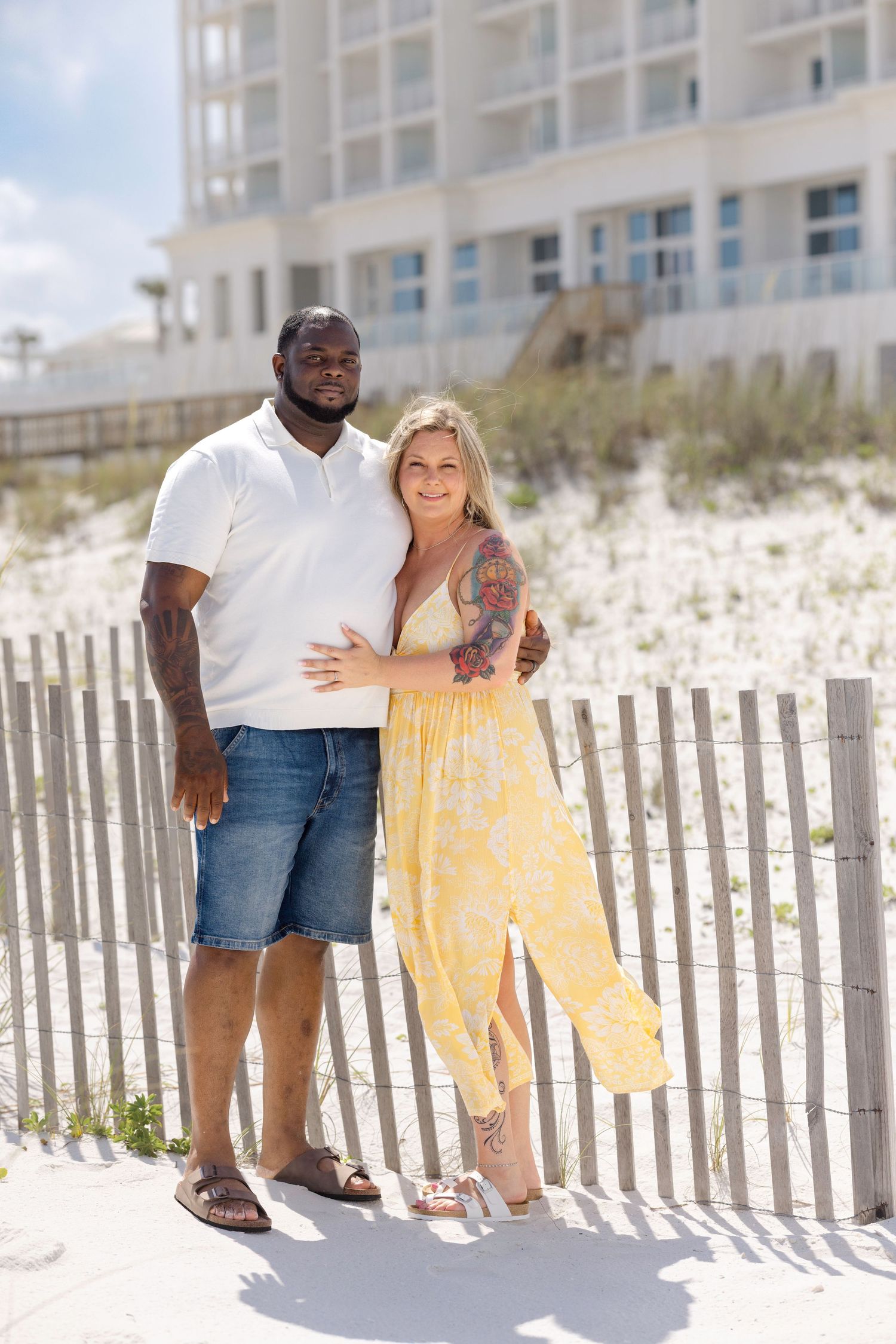 Interracial couple posing together on a sandy beach near a wooden fence, man in white polo and woman in yellow dress.