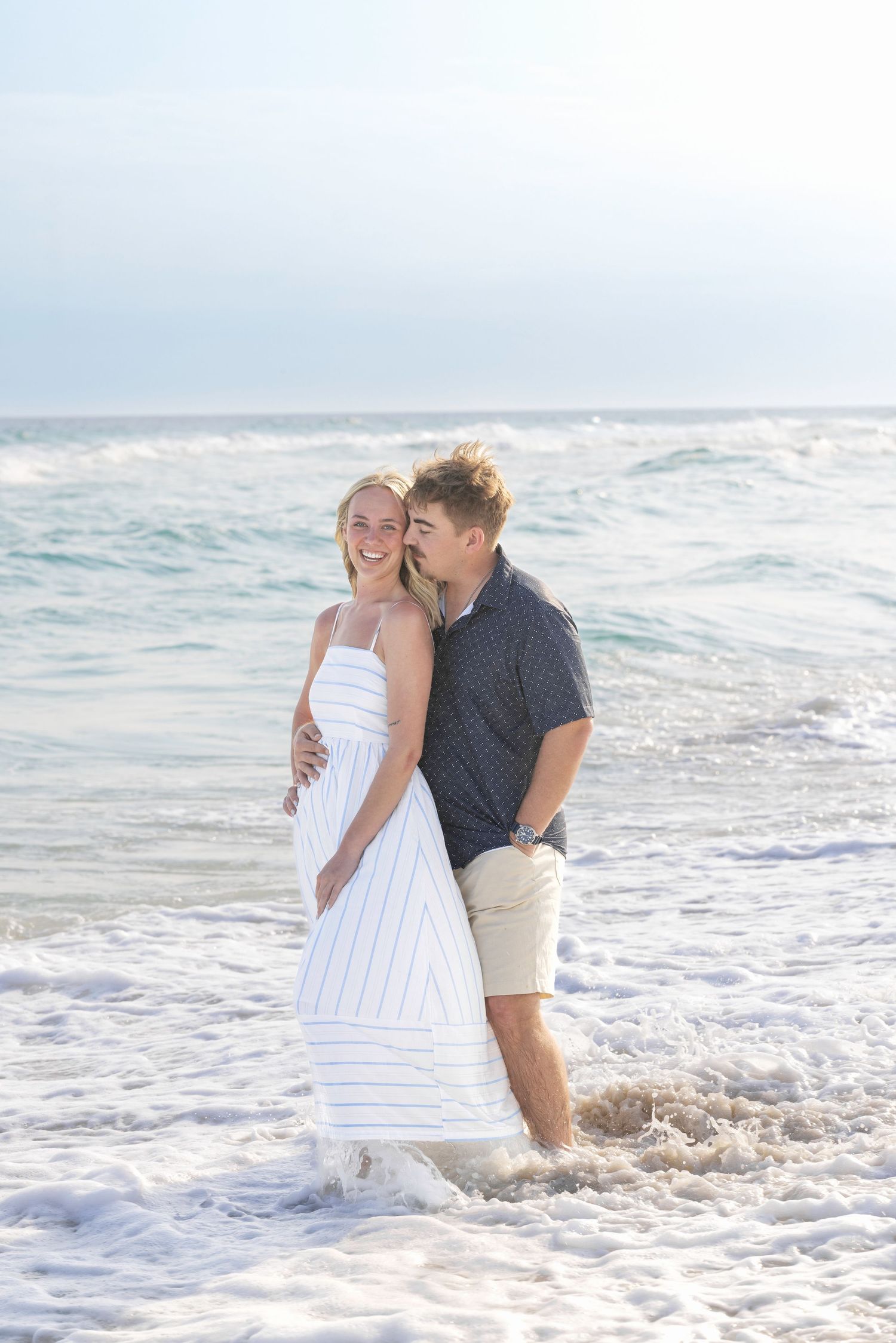 A happy couple stands in ocean waves on a sunny beach, the woman in a white dress laughing as the man kisses her cheek.