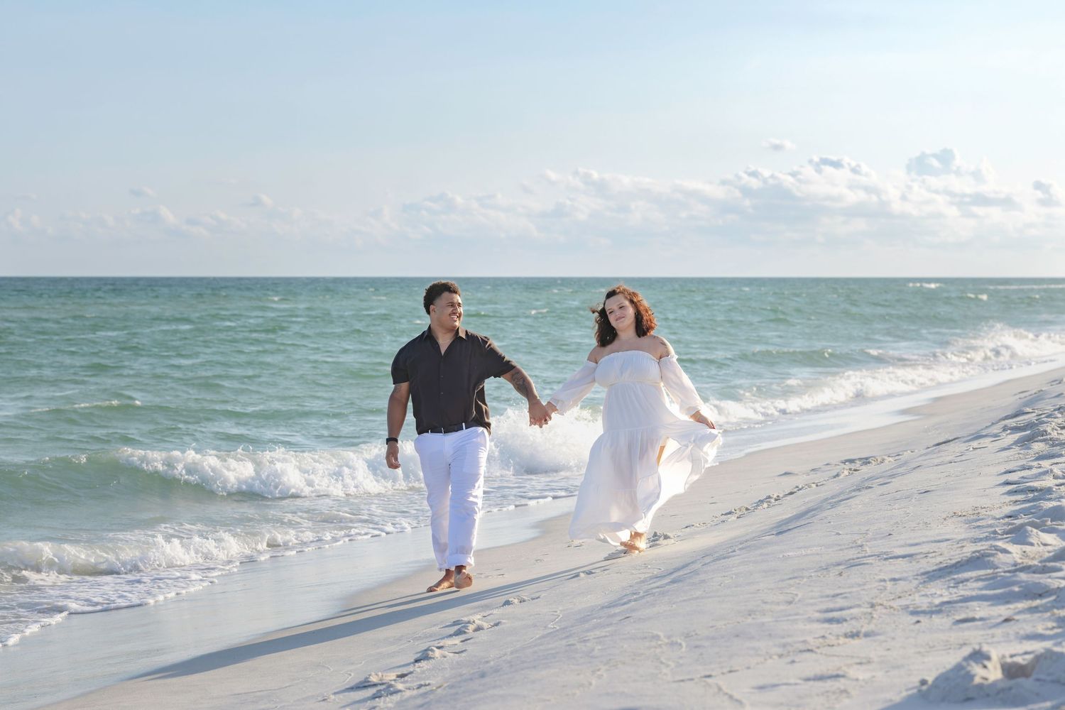 A couple walks hand-in-hand on a white sand beach, waves gently crashing behind them on a sunny day.