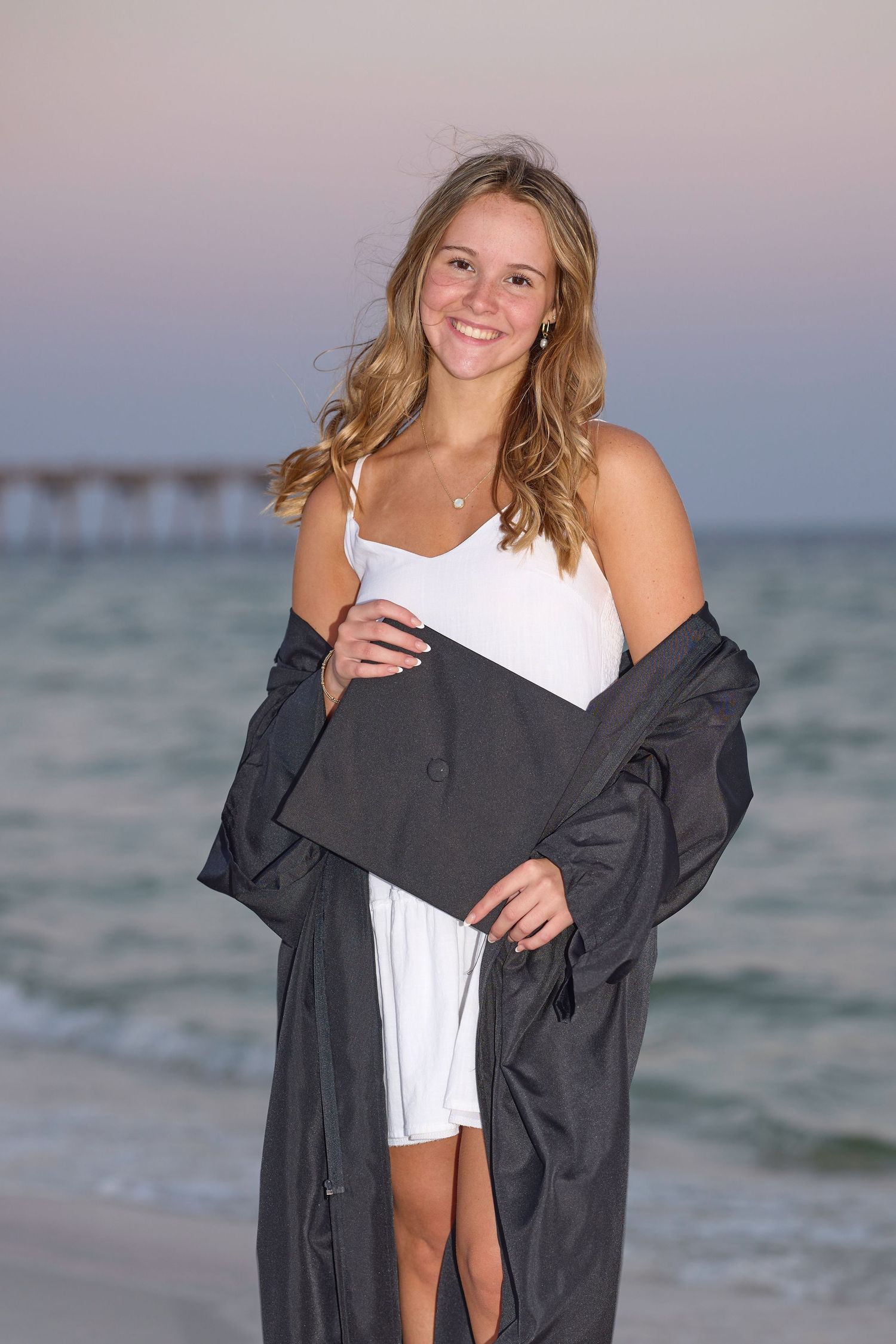 Smiling graduate poses on beach at sunset wearing white dress holding black cap and gown.