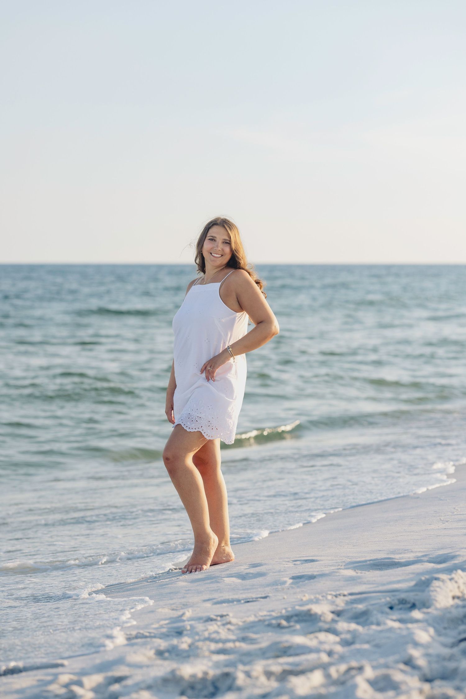 A smiling young woman in a white sundress stands barefoot on a sunny white sand beach with calm ocean waves behind her.