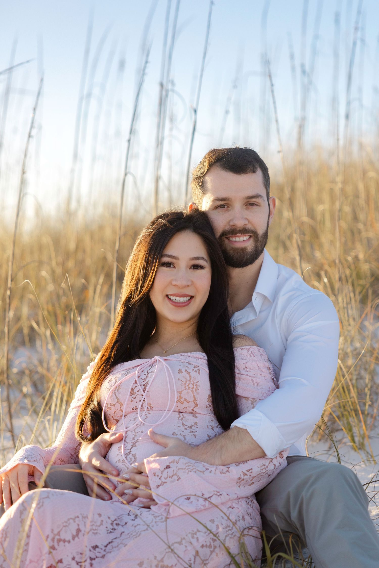A couple sits together on a beach with sand dunes and sea grass in the background during golden hour.