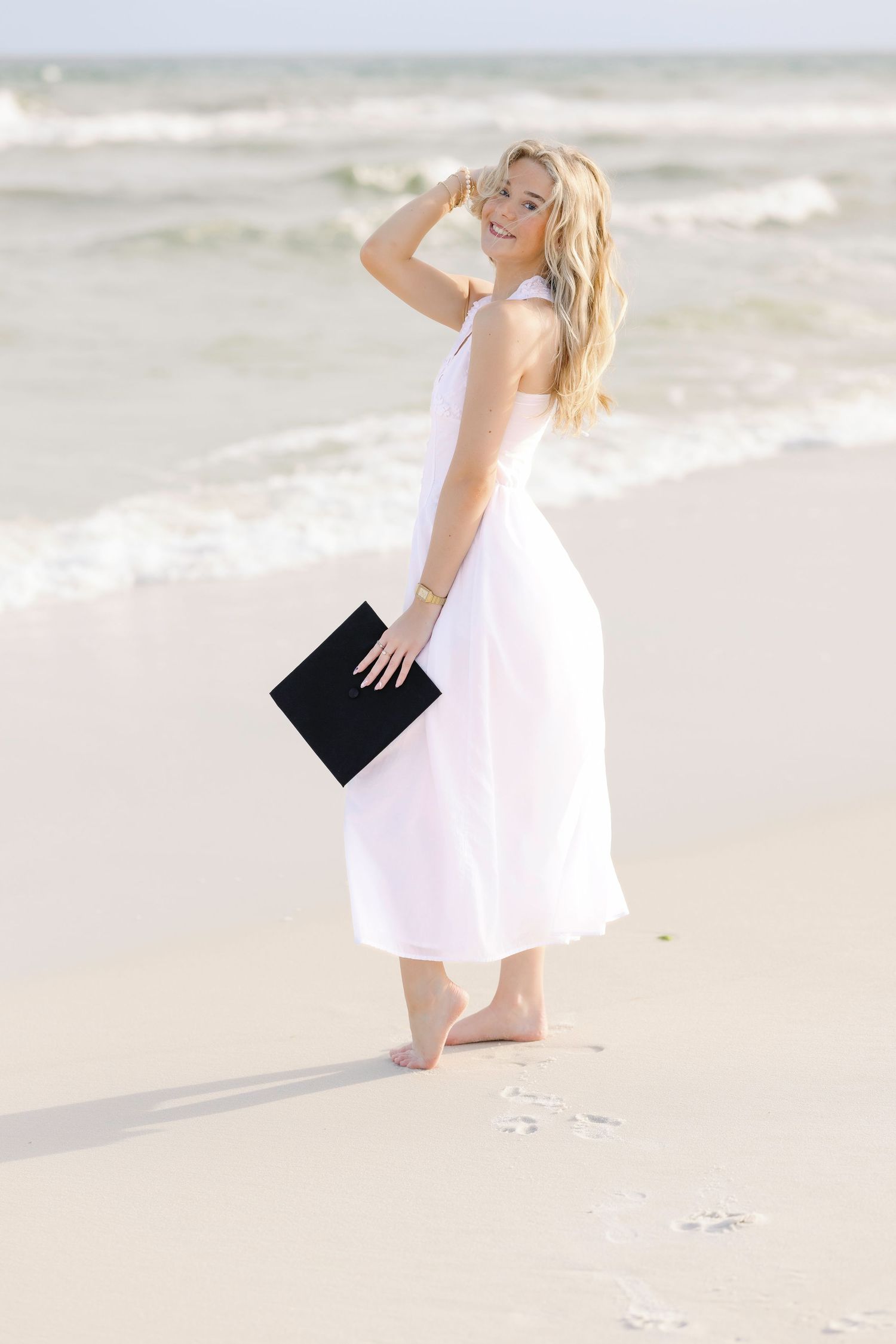 A person in a flowing white dress holds a black book while walking along a serene beach at sunset.