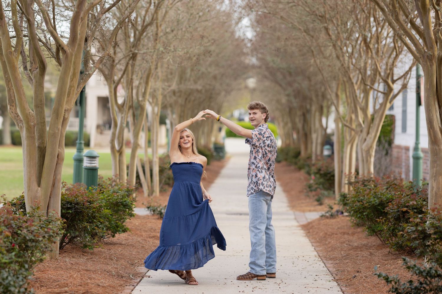 A couple dances together on a tree-lined pathway with the woman twirling in a flowing blue dress.