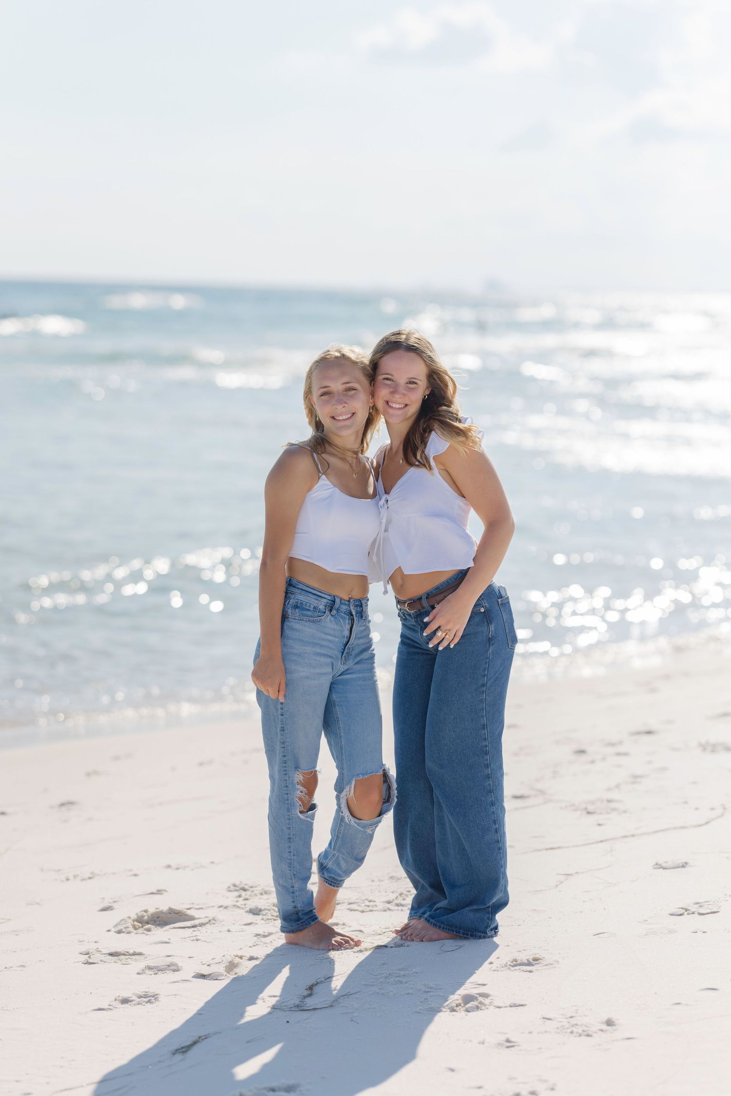 Two friends in white crop tops and denim jeans pose together on a sunny beach with sparkling ocean waves in background.