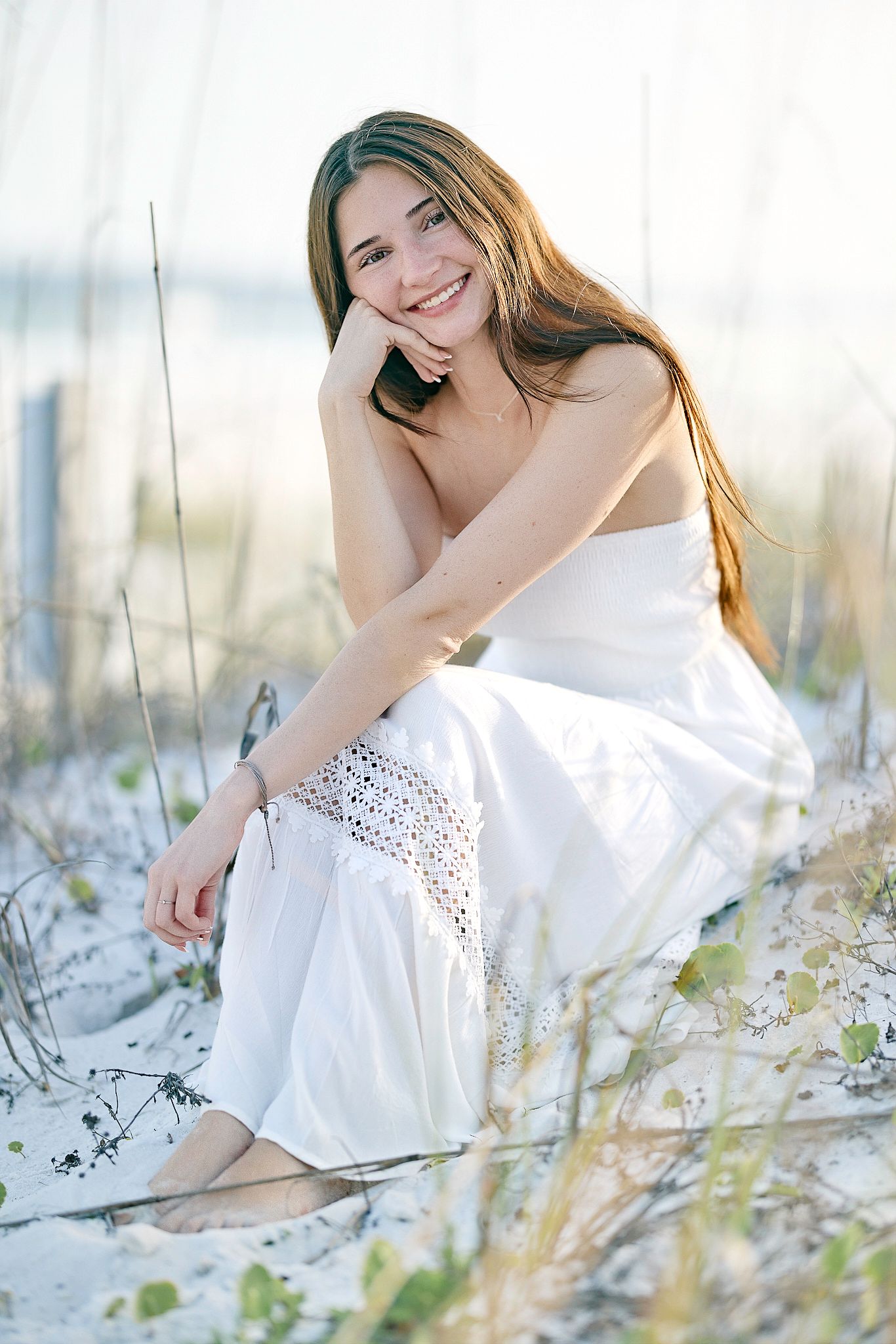 Candid beach portrait sitting in white dress among coastal vegetation.