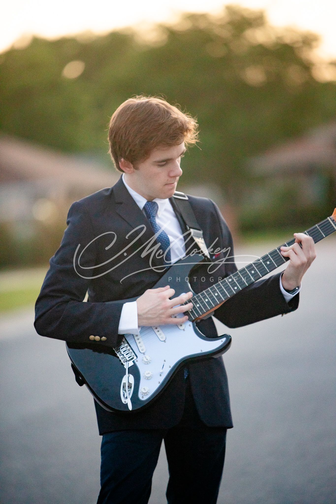 A musician in a formal black suit plays an electric Stratocaster guitar during golden hour outdoors.
