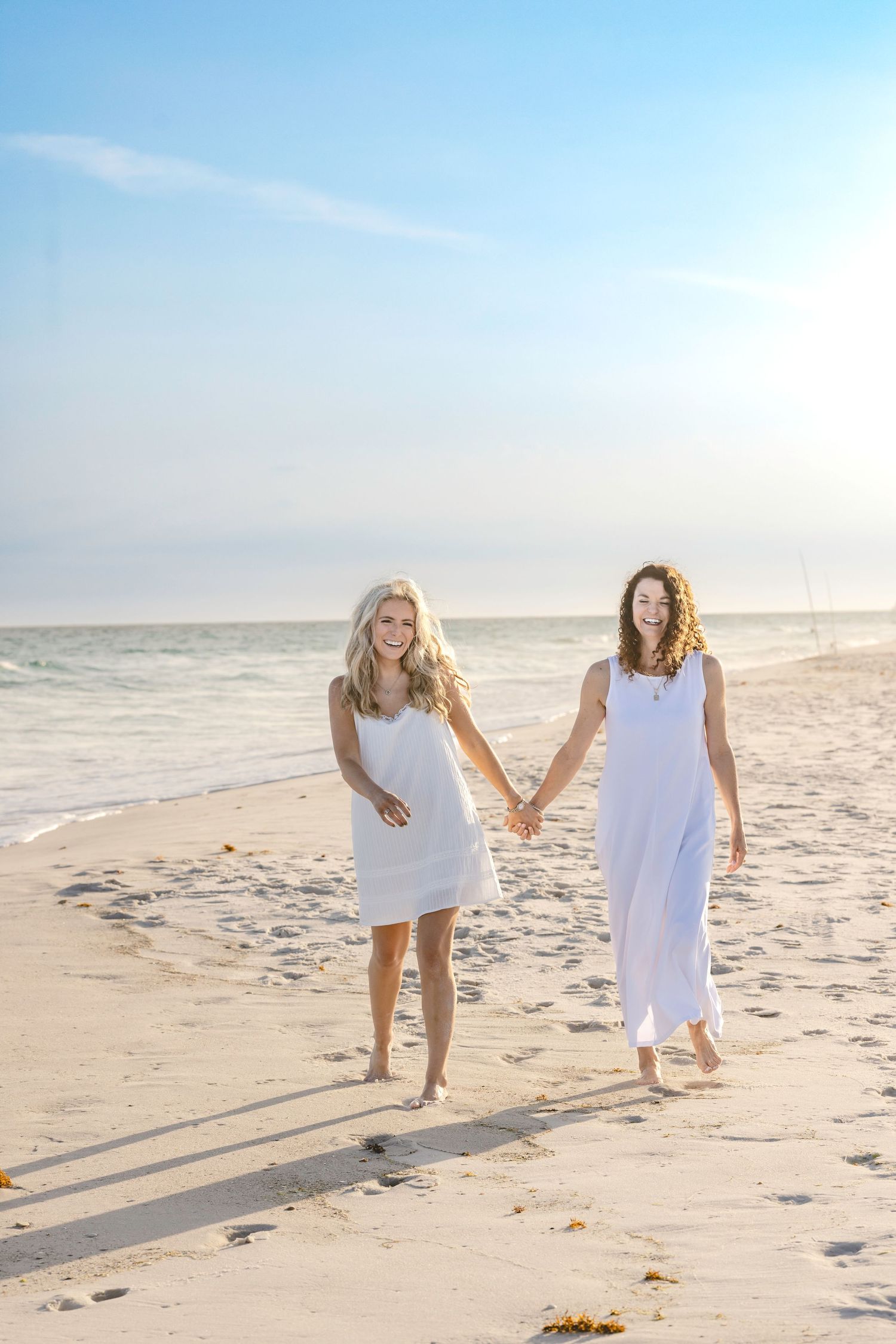 Two women in white dresses holding hands and walking barefoot on a sunny beach with ocean waves in the background.
