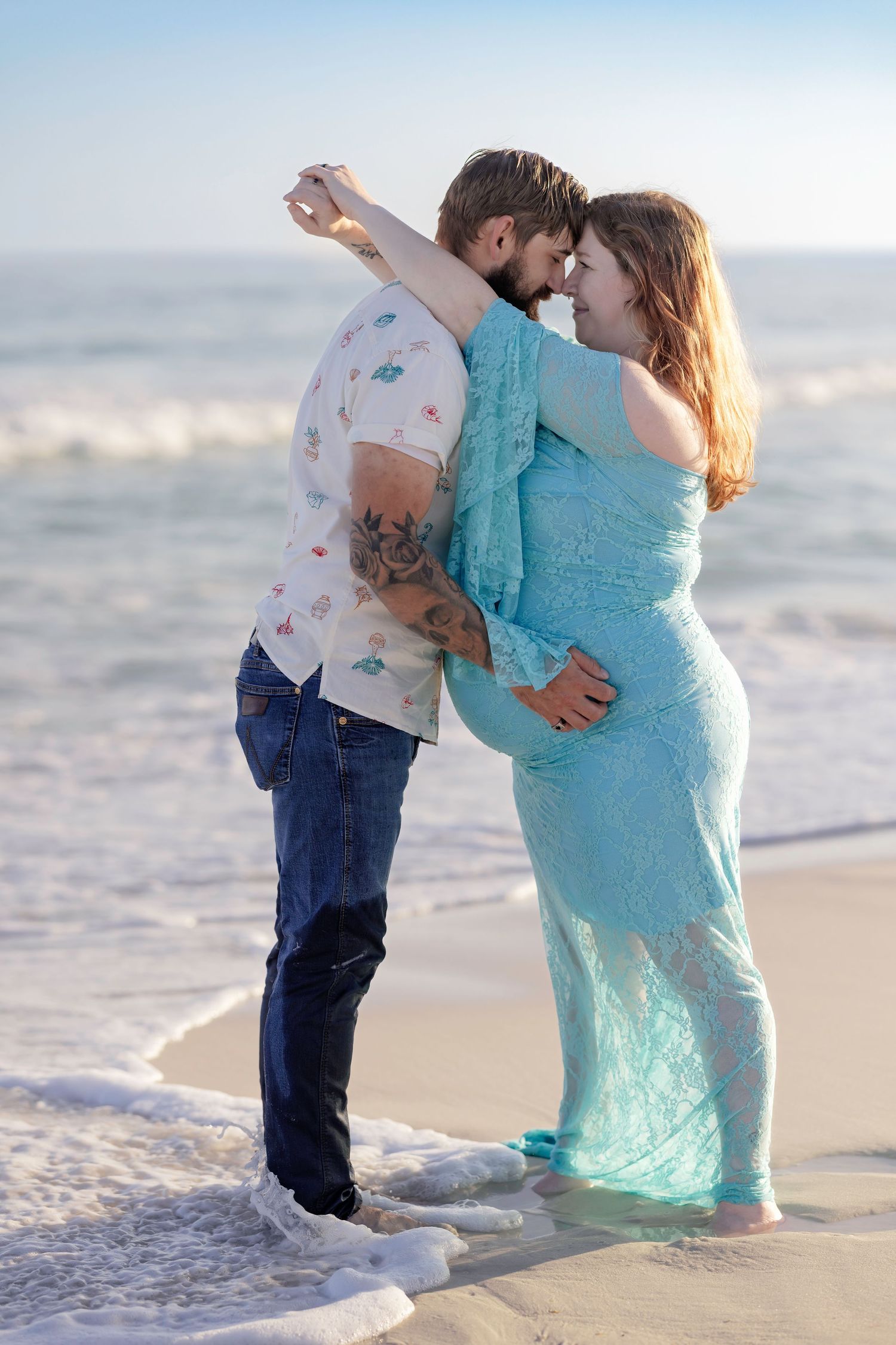 Expecting couple dancing romantically at ocean's edge, woman in turquoise maternity gown.