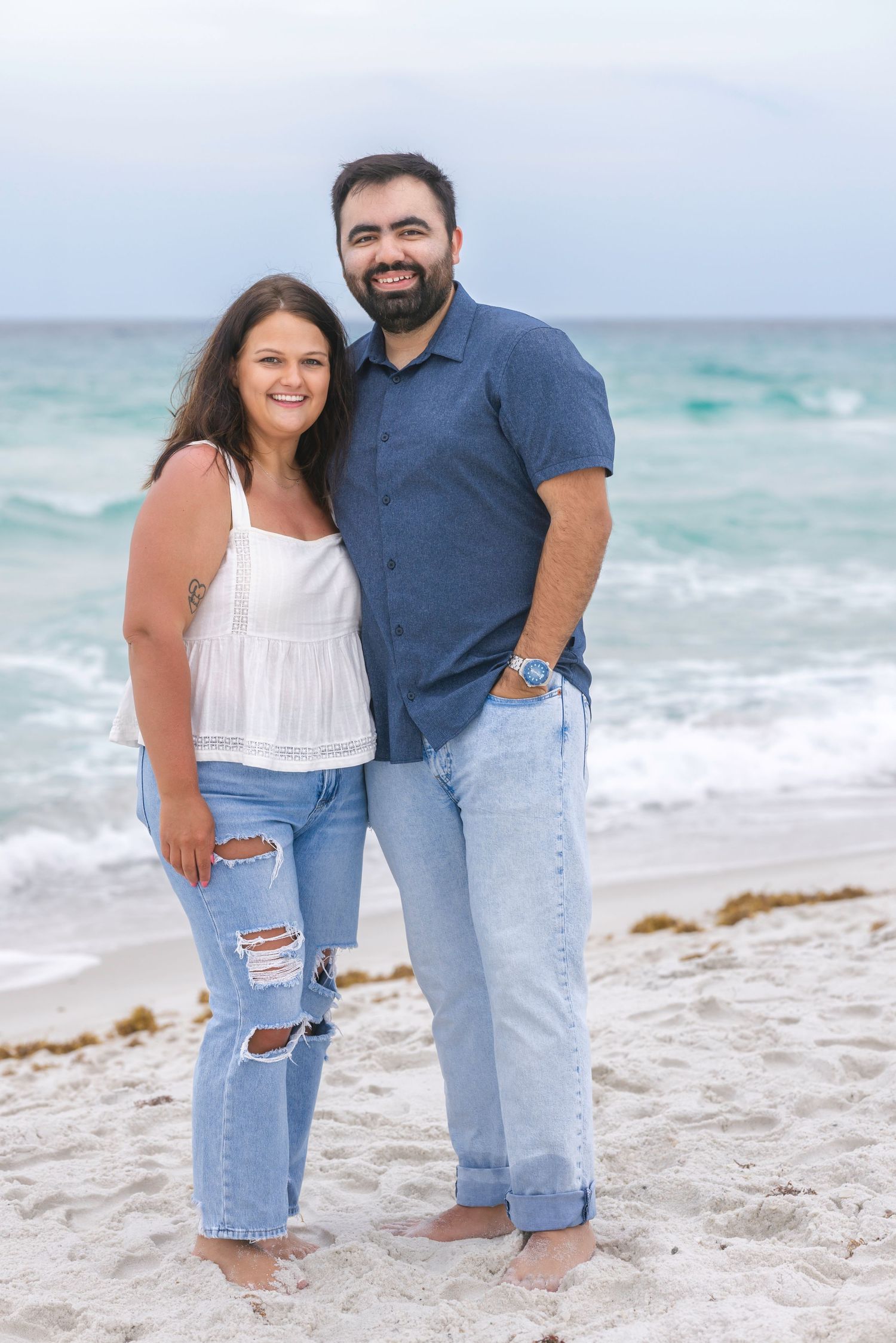 A smiling couple poses barefoot on a white sand beach with turquoise ocean waves behind them.
