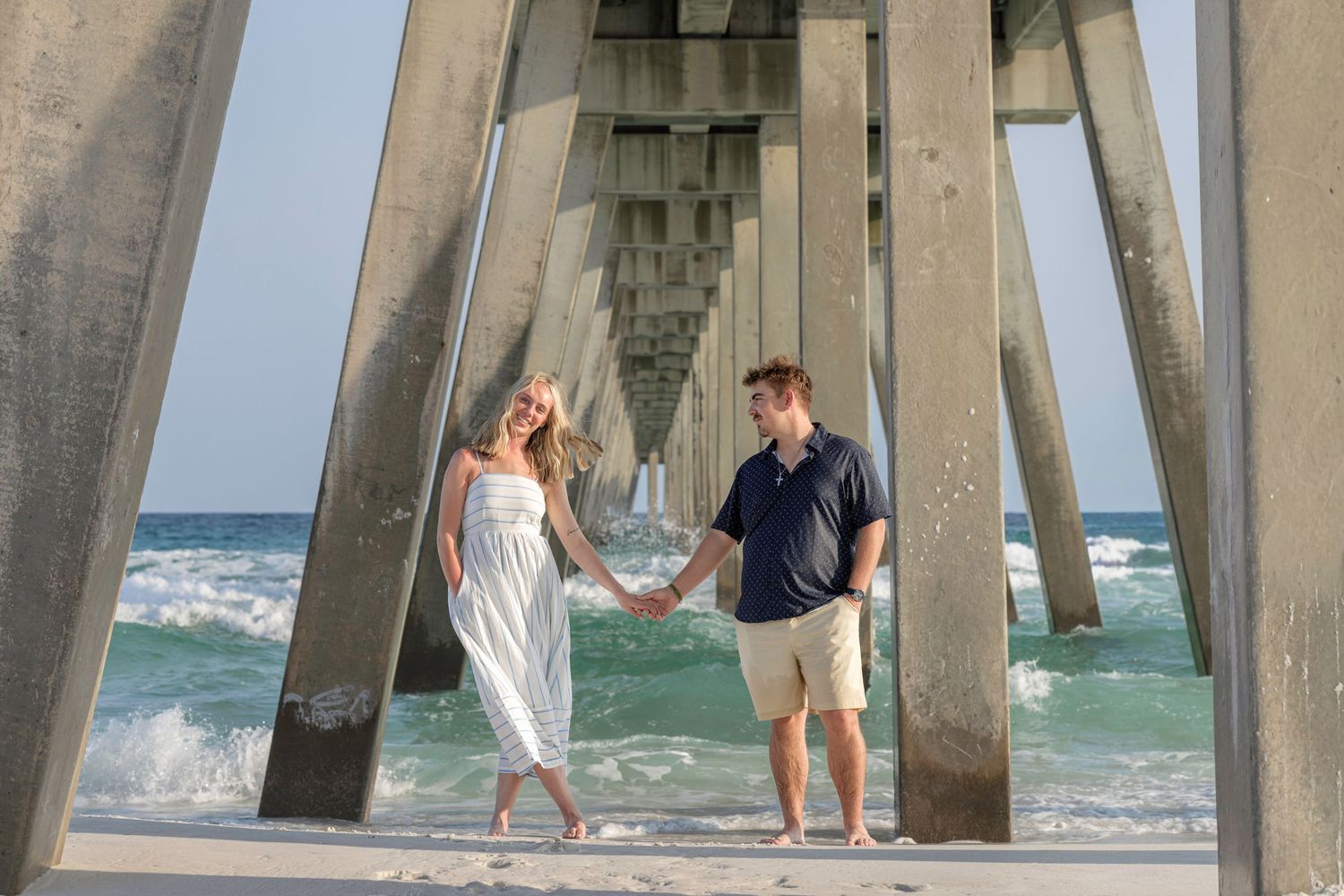 Couple holding hands under a beach pier with ocean waves and turquoise water in the background.