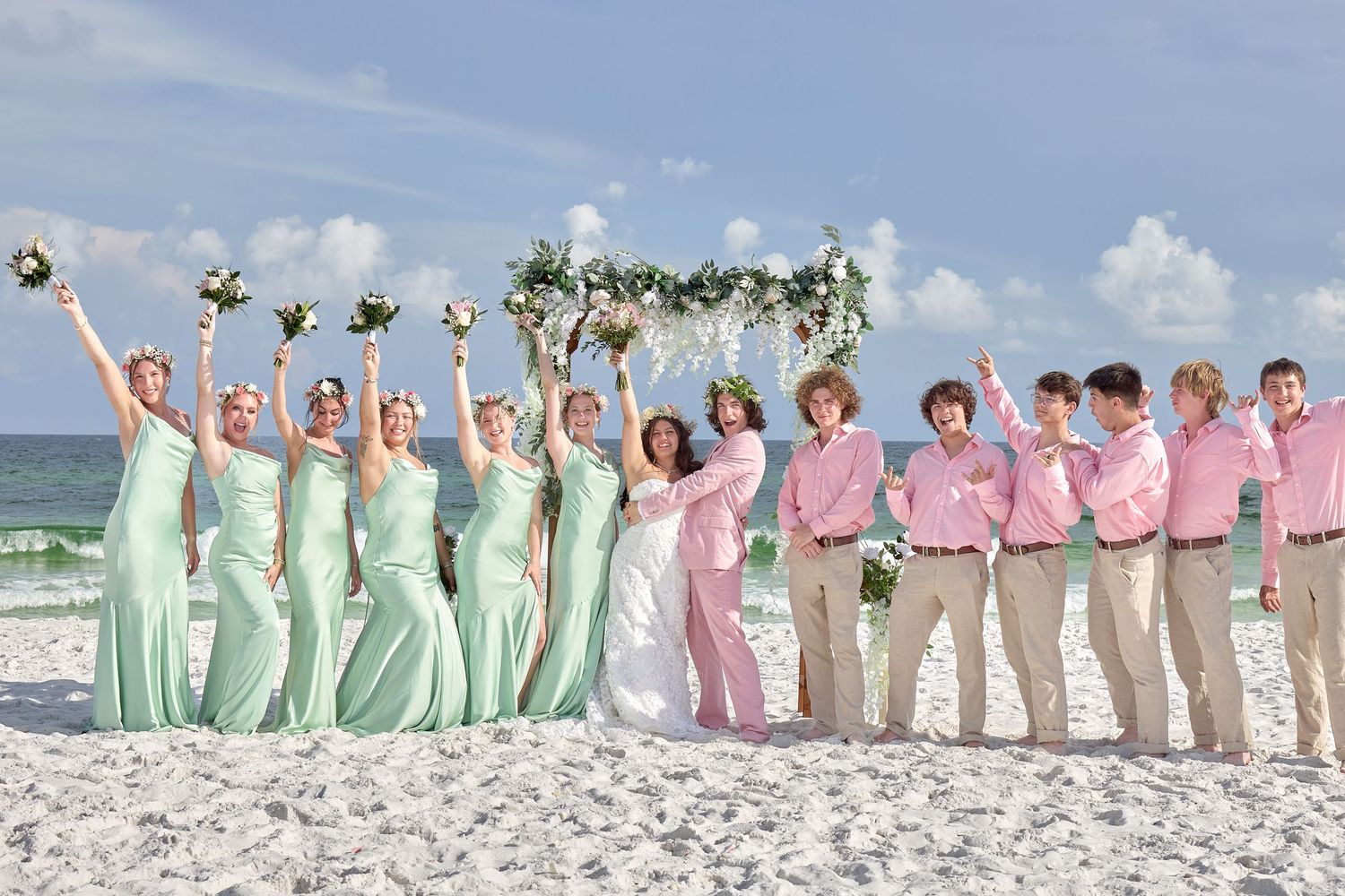 Wedding party poses on beach, bridesmaids in mint gowns, groomsmen in pink shirts, celebrating together.