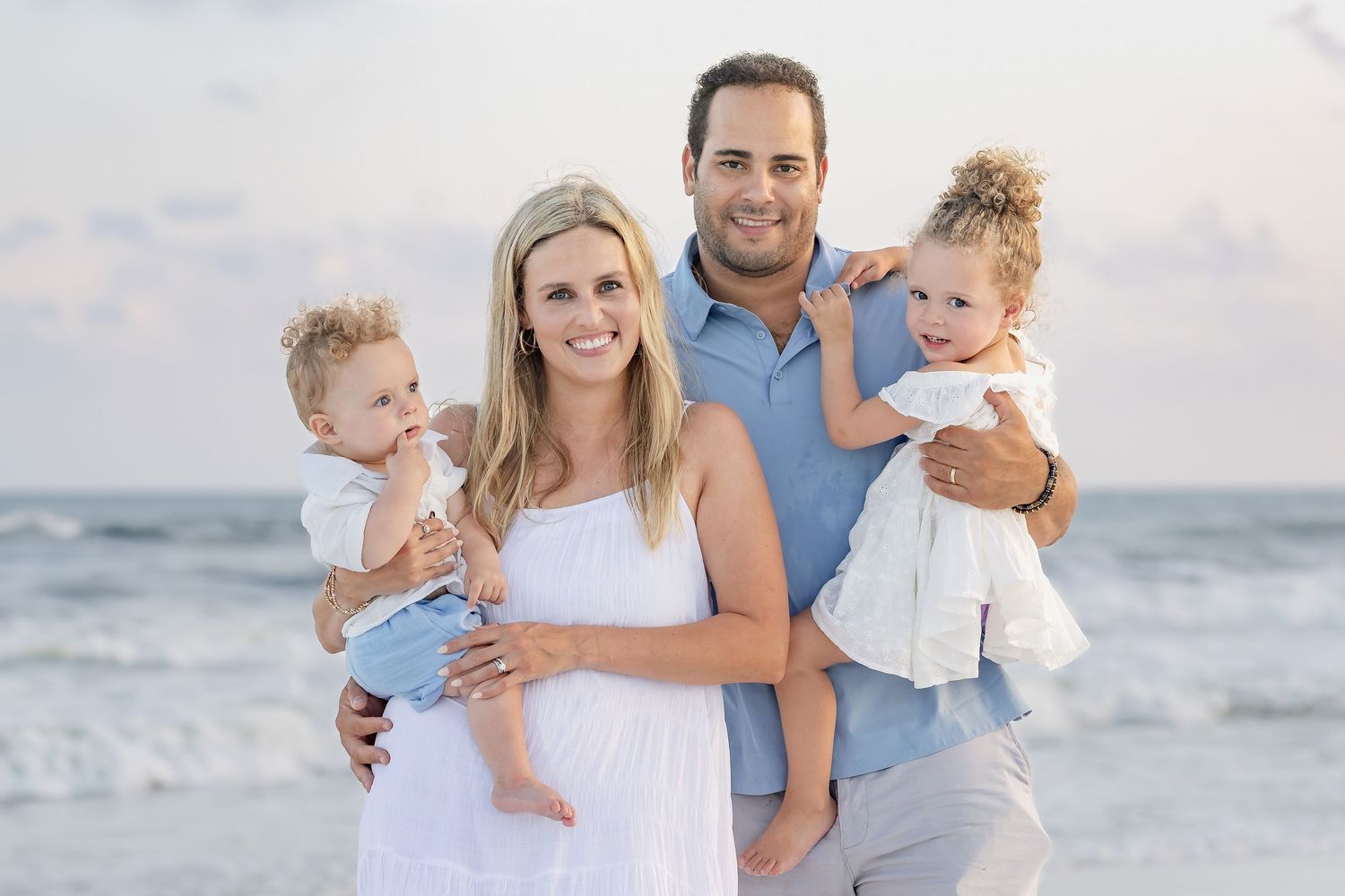 Happy family of four posing on a beach, parents holding two young children, with ocean waves in the background.