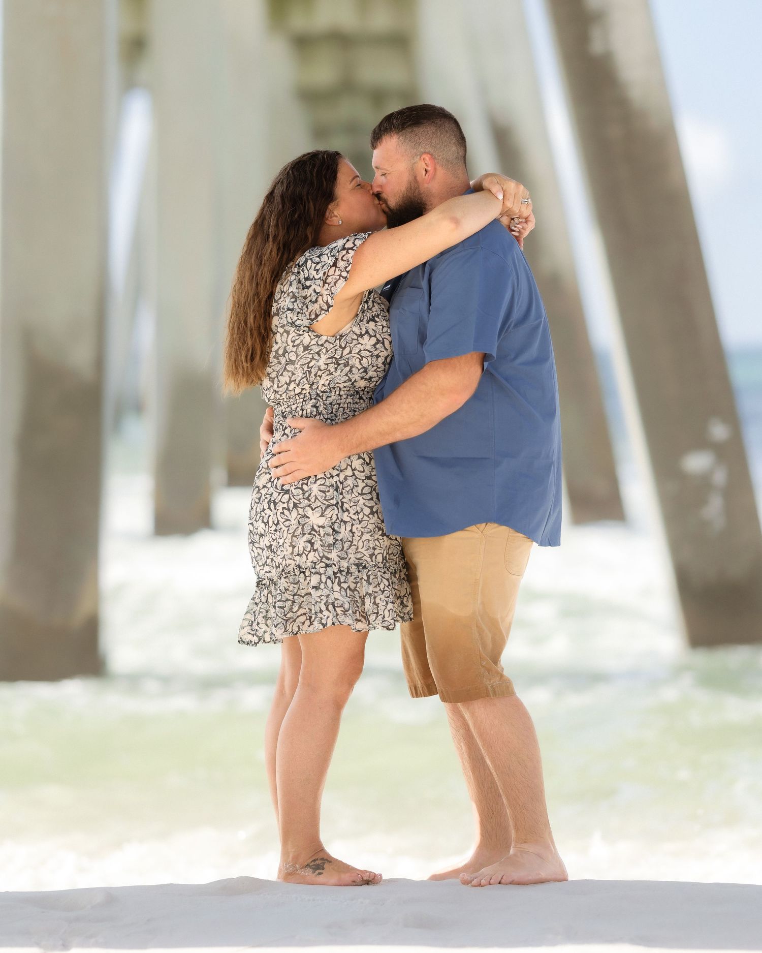 Couple kissing on beach under pier in romantic seaside engagement photo.