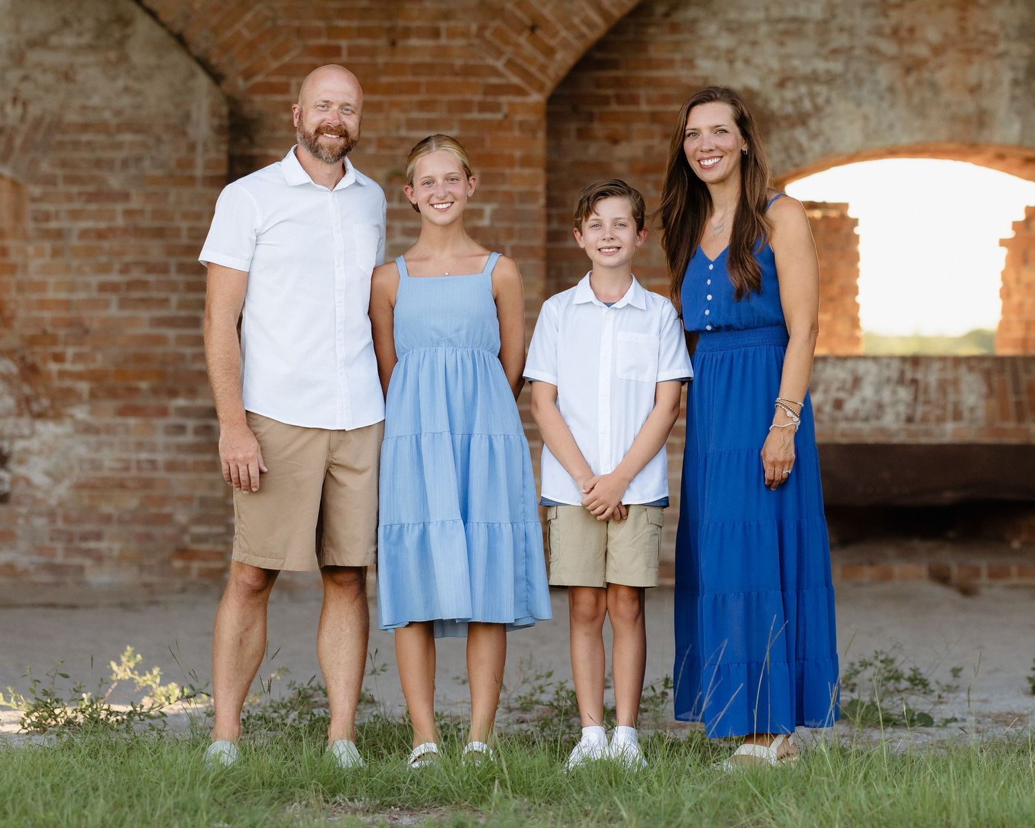 Family of four posing outdoors near rustic brick arches, wearing coordinating blue and white outfits on a sunny day.