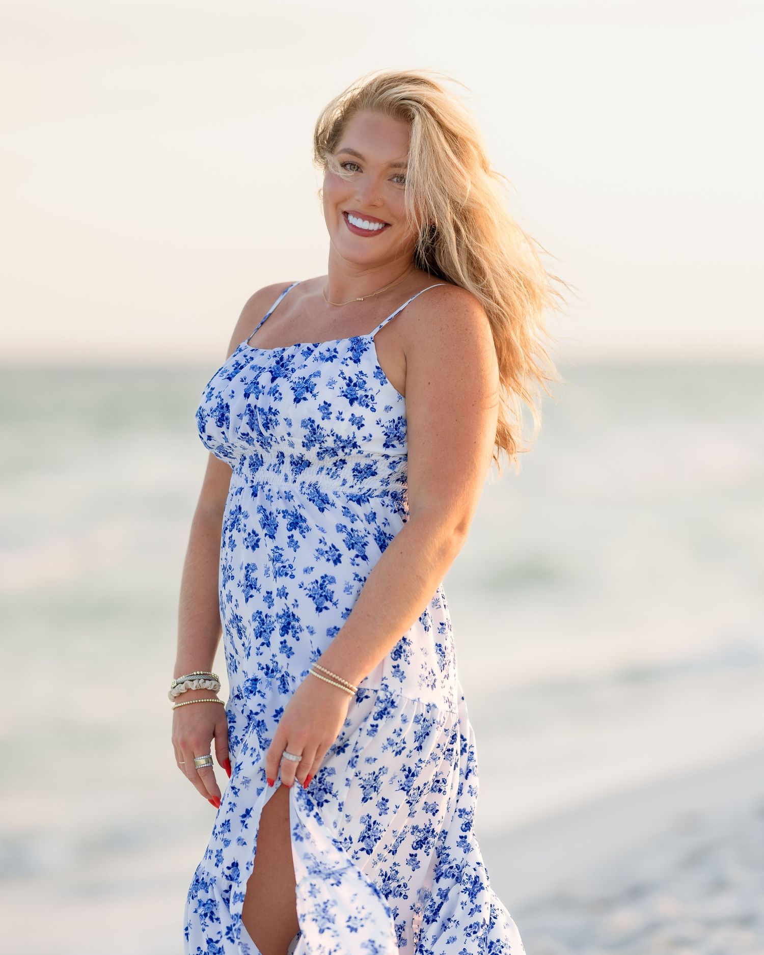Smiling blonde woman in blue floral sundress posing on a sunny beach with ocean waves in the background.