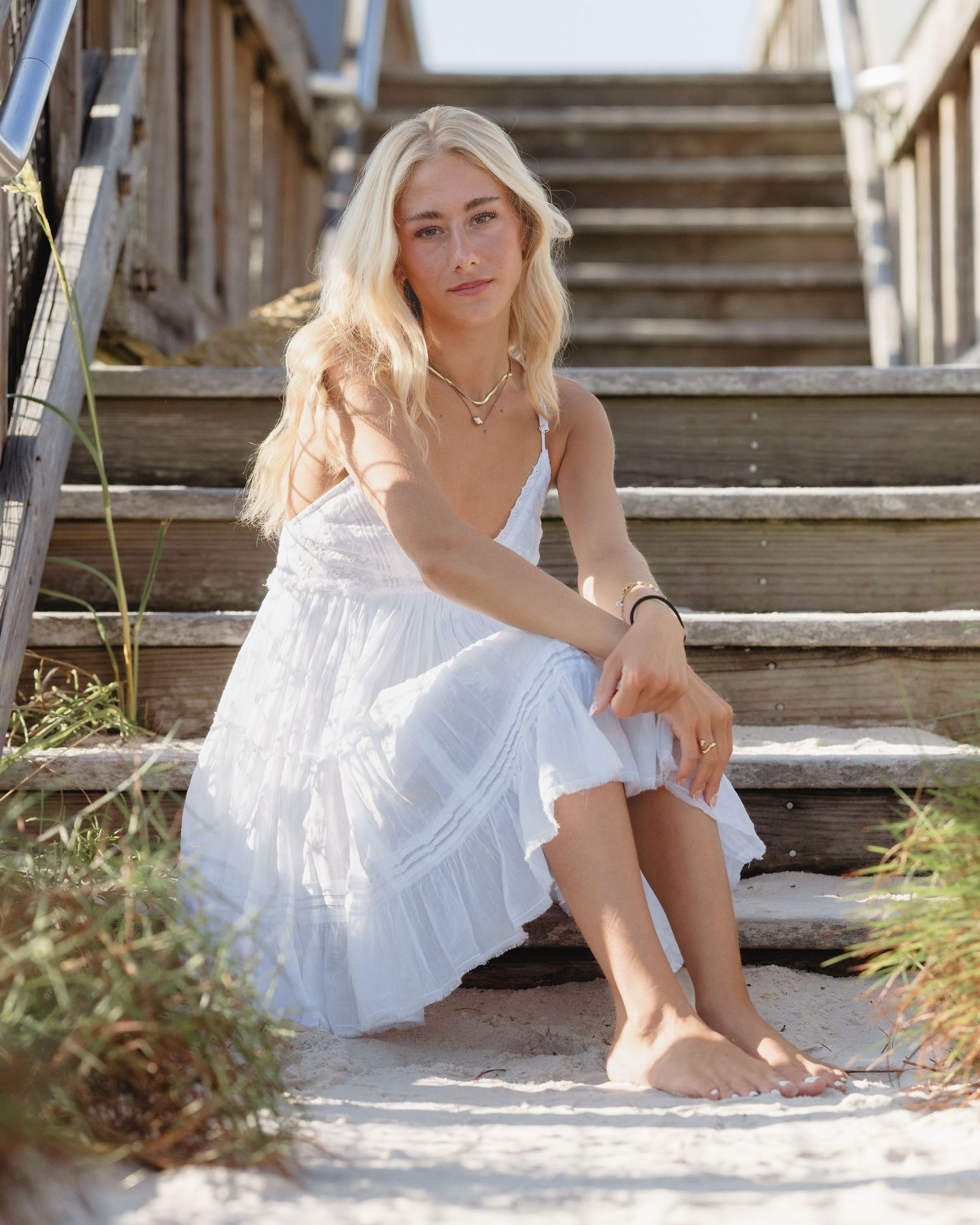 Blonde woman in white sundress sitting on weathered beach stairs surrounded by sand dunes.