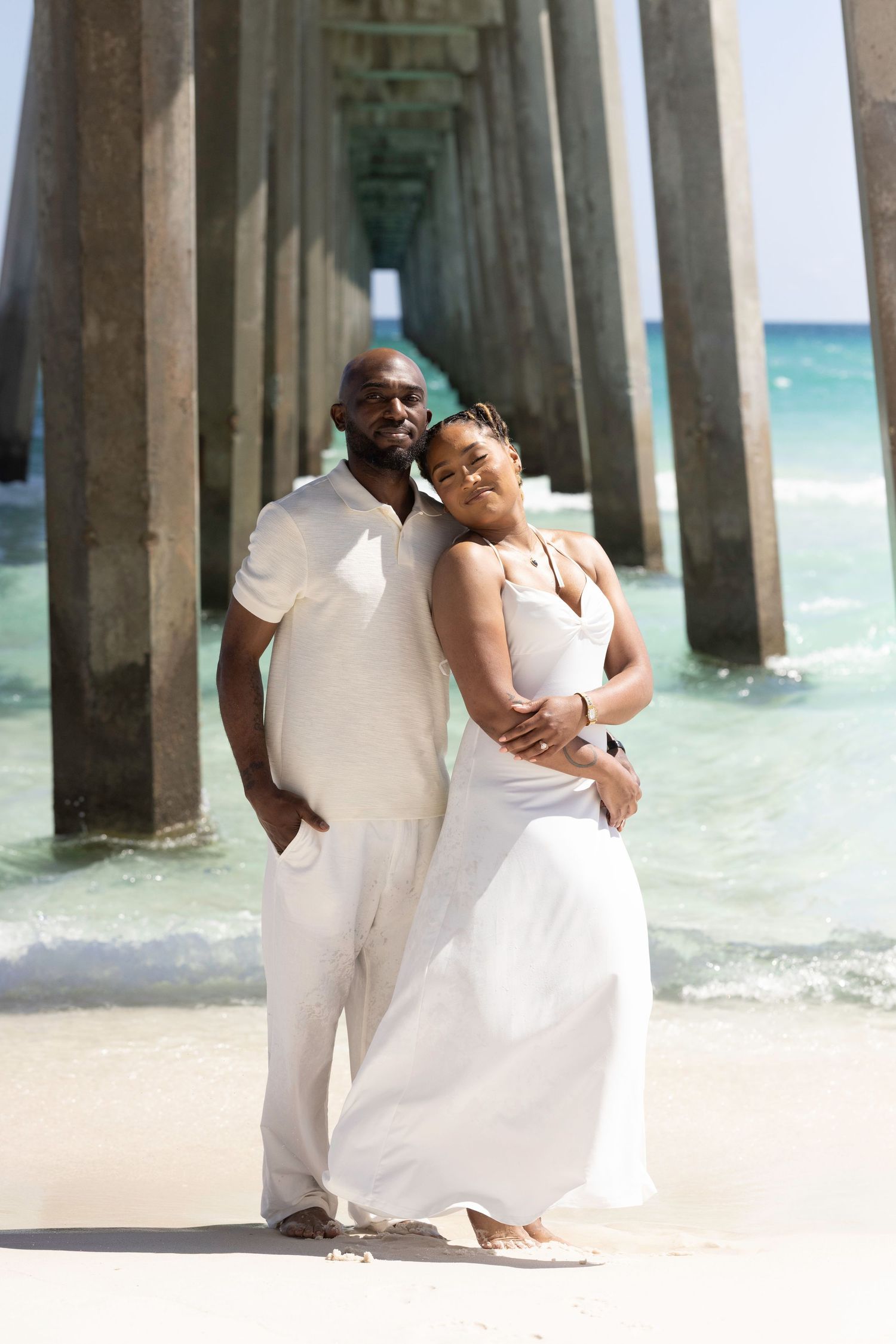 Couple in white outfits posing under a beach pier with turquoise ocean water in the background.
